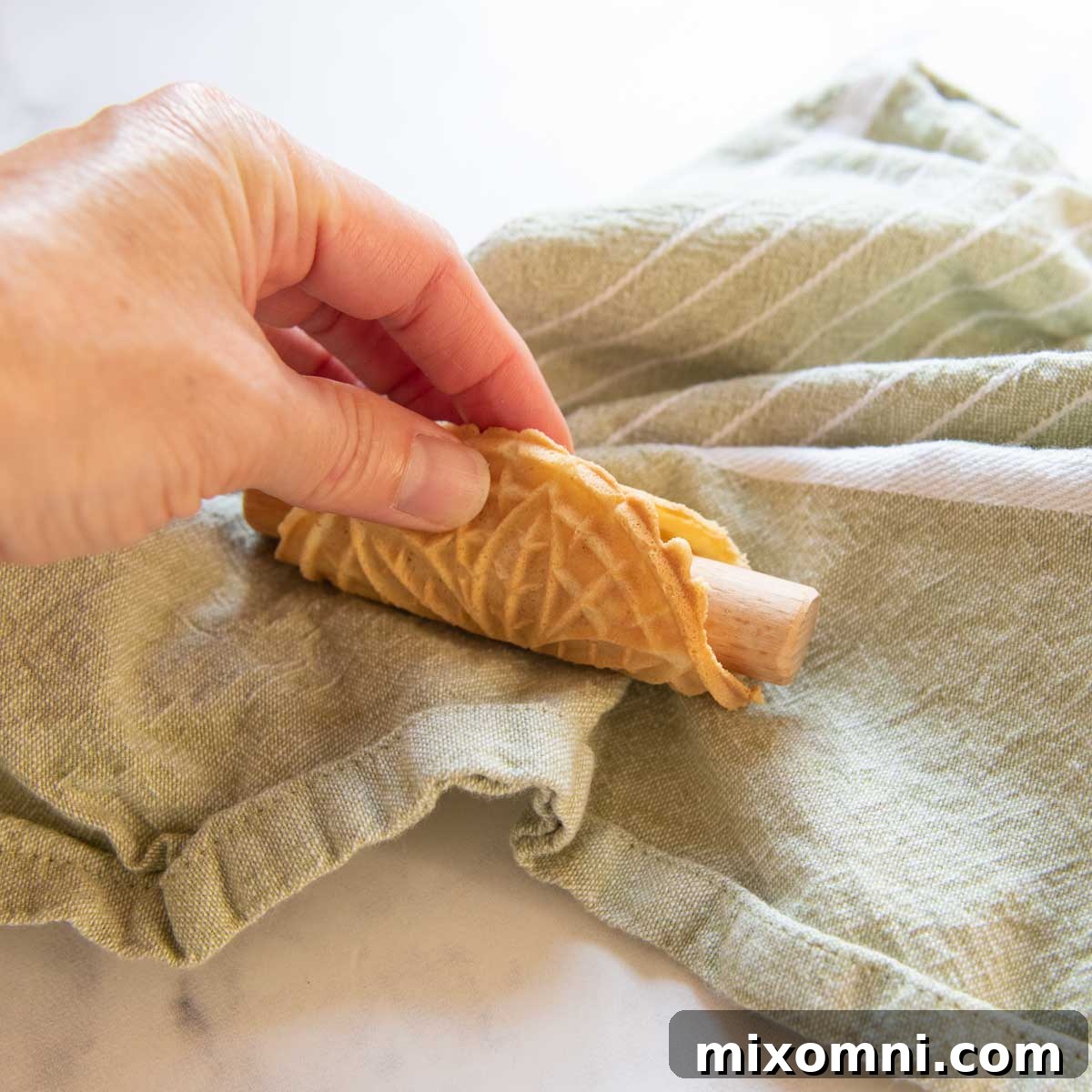 A small, warm pizzelle being carefully wrapped around a rod to create a cannoli shape.