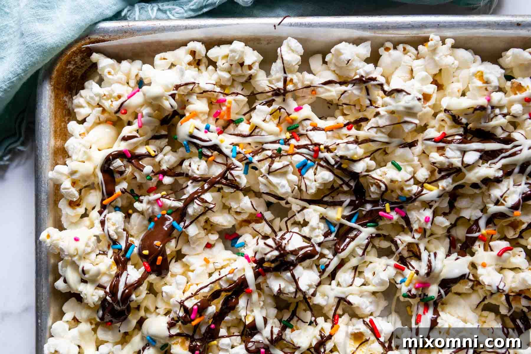 An overhead shot of a baking sheet filled with popcorn, artfully drizzled with chocolate and adorned with colorful sprinkles.