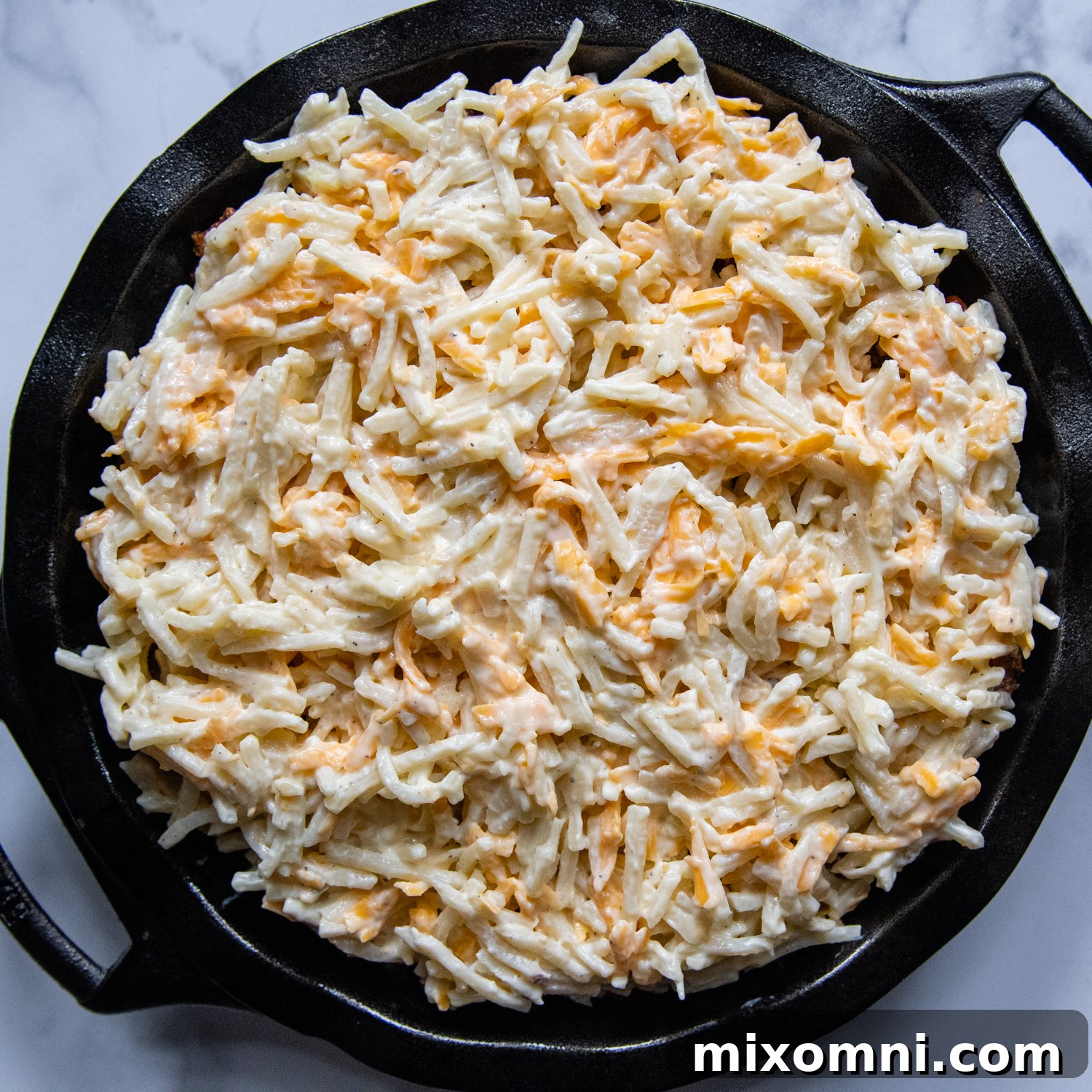 The cheesy hash brown topping spread over the savory meat filling in a baking dish, ready for the oven.