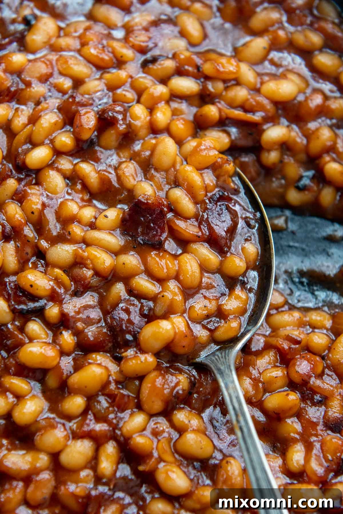 An antique spoon scoops a generous portion of southern baked beans from a rustic bowl, emphasizing their rich, saucy texture.