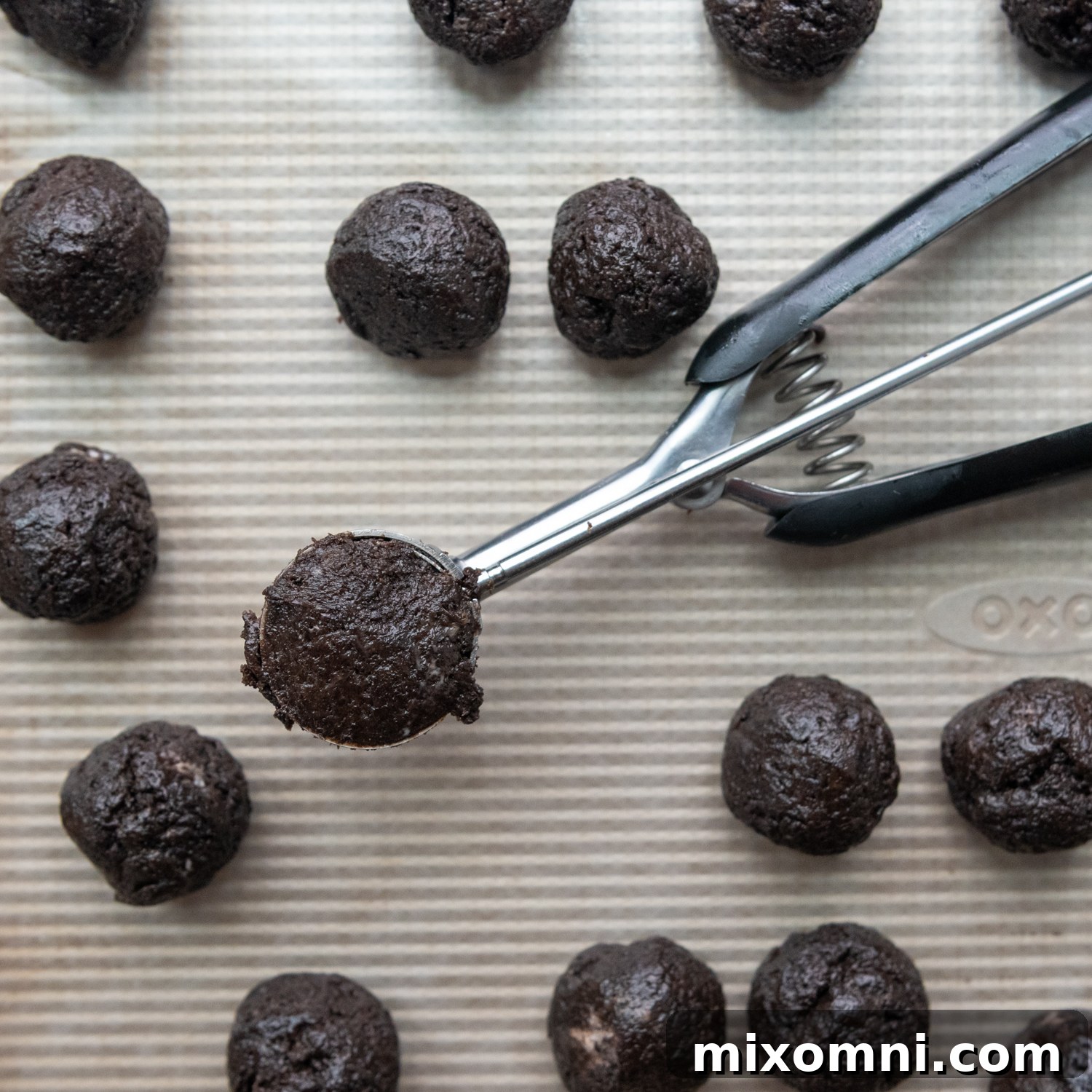 A small cookie scoop precisely dividing the gluten-free Oreo truffle mixture onto a lined baking sheet for consistent sizing.