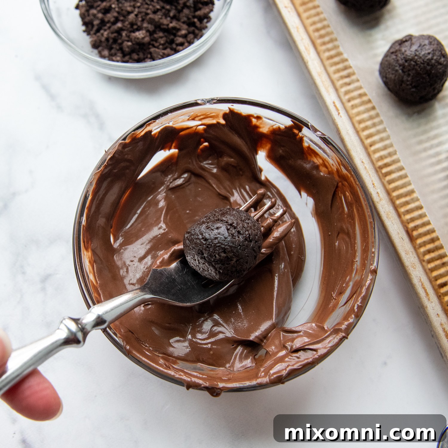 A gluten-free Oreo truffle being carefully lifted out of a bowl of melted chocolate with a fork, showing excess chocolate dripping away for a smooth finish.