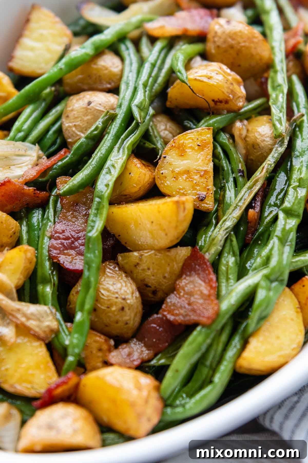 A close-up shot of crispy roasted green beans and potatoes with bacon on a baking sheet, glistening with flavor.