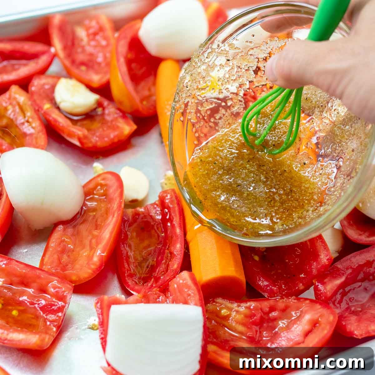 A small bowl of olive oil being poured over fresh tomatoes, carrots, and onion chunks on a sheet pan.