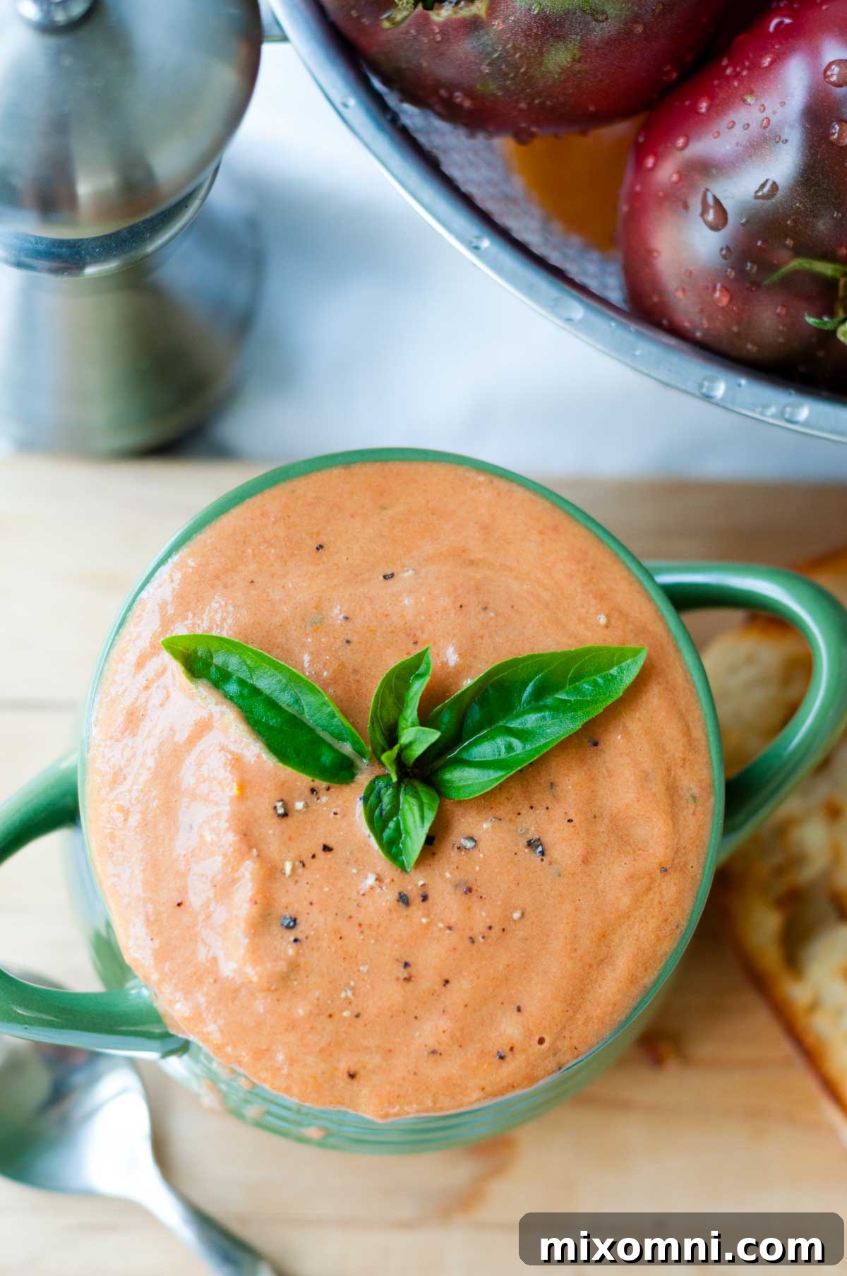 A top-down view of a serving of creamy roasted tomato soup, with fresh tomatoes and basil next to it.