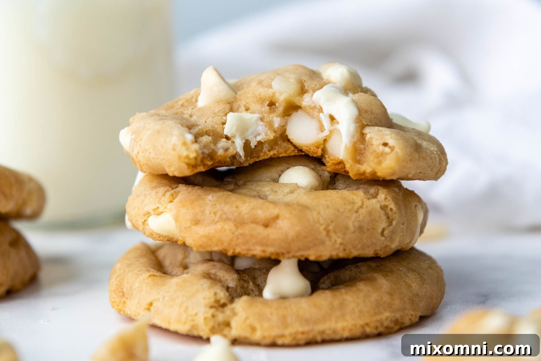 A stack of three perfectly baked gluten-free white chocolate macadamia nut cookies, with a tempting bite taken out of the top one, showcasing its tender texture.