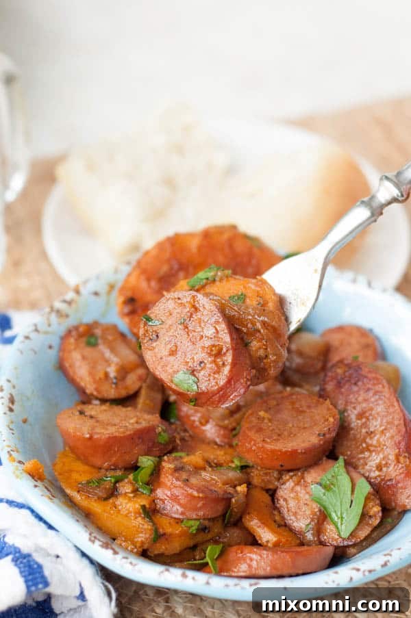A close-up shot of a fork lifting a piece of smoked kielbasa and sweet potato from a rich red goulash in a blue bowl, highlighting the tender textures and vibrant colors.