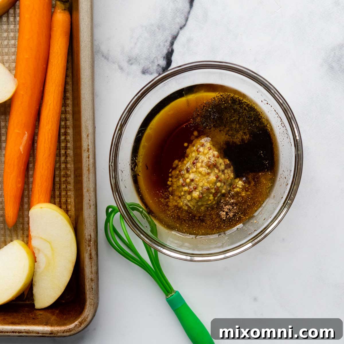 Close-up of the maple mustard glaze being drizzled over cut carrots and apples on a baking sheet.