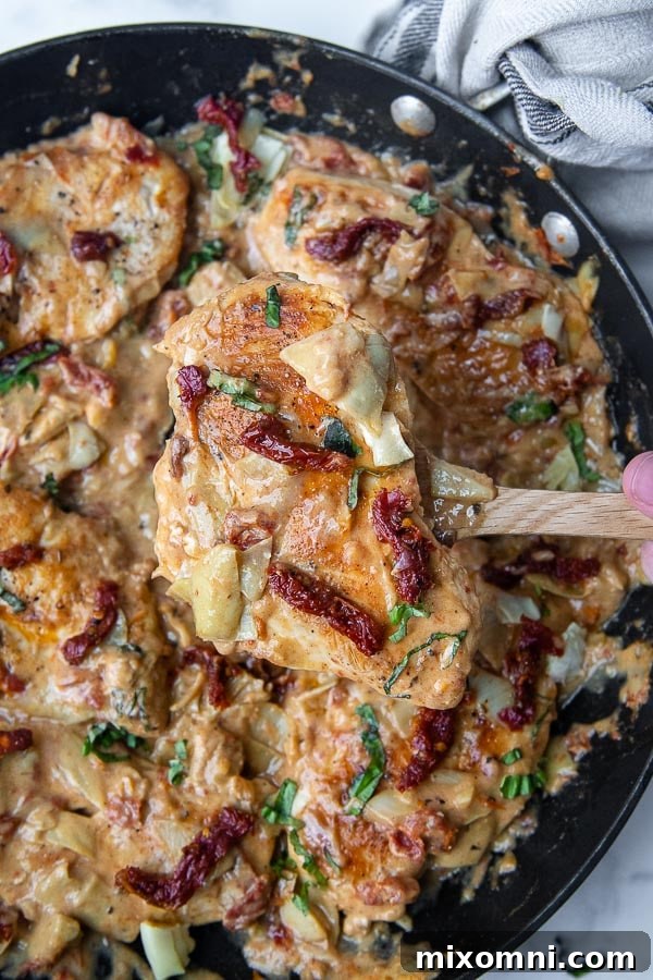 A wooden spoon lifting a piece of chicken from a pan of creamy sun-dried tomato and artichoke sauce.