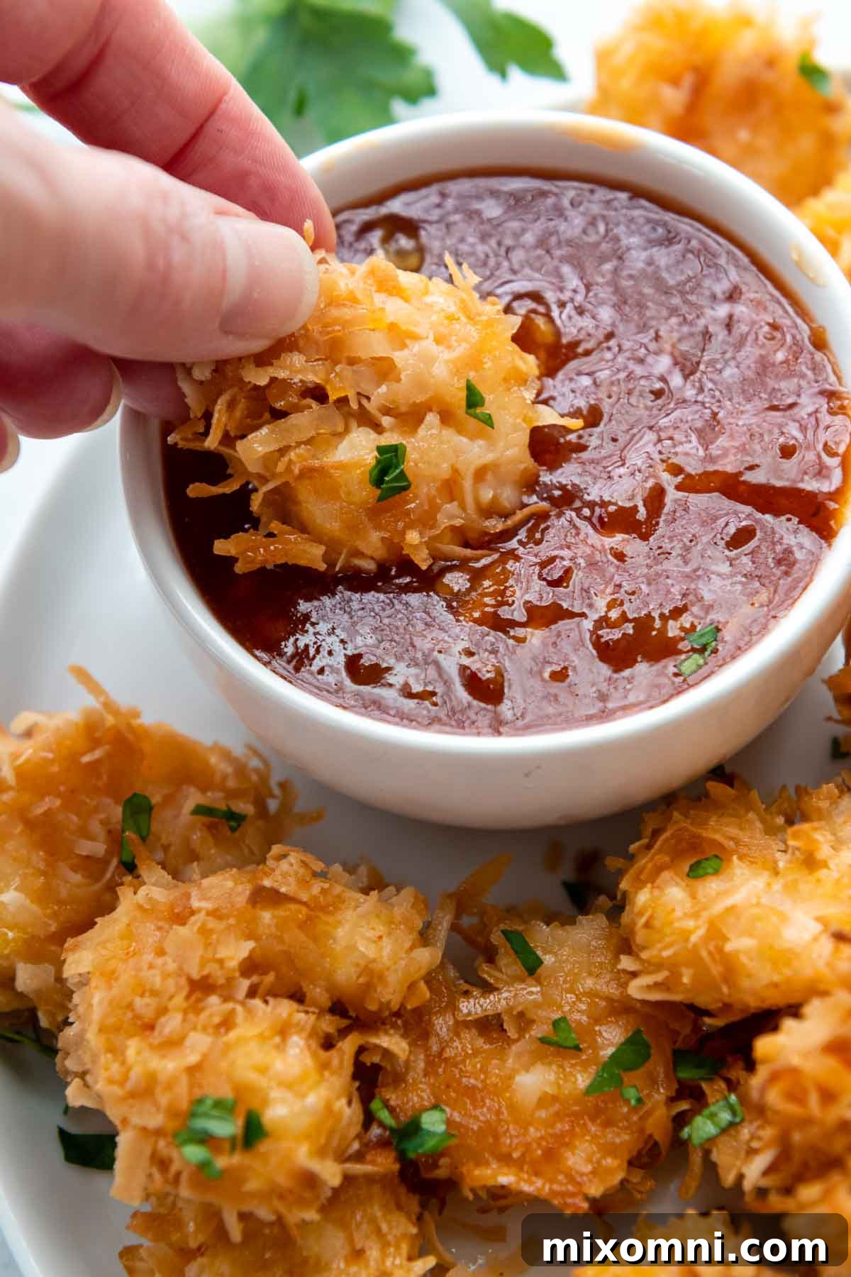 A succulent gluten-free coconut shrimp being dipped into a luscious, sweet and spicy sauce, ready to be enjoyed.