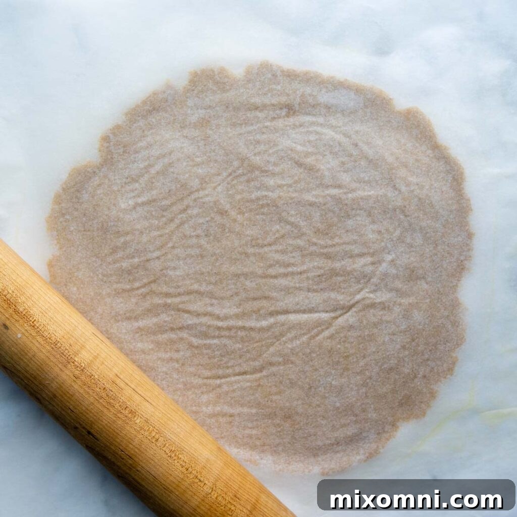 Almond flour pizza crust being rolled out evenly between two sheets of parchment paper using a rolling pin.