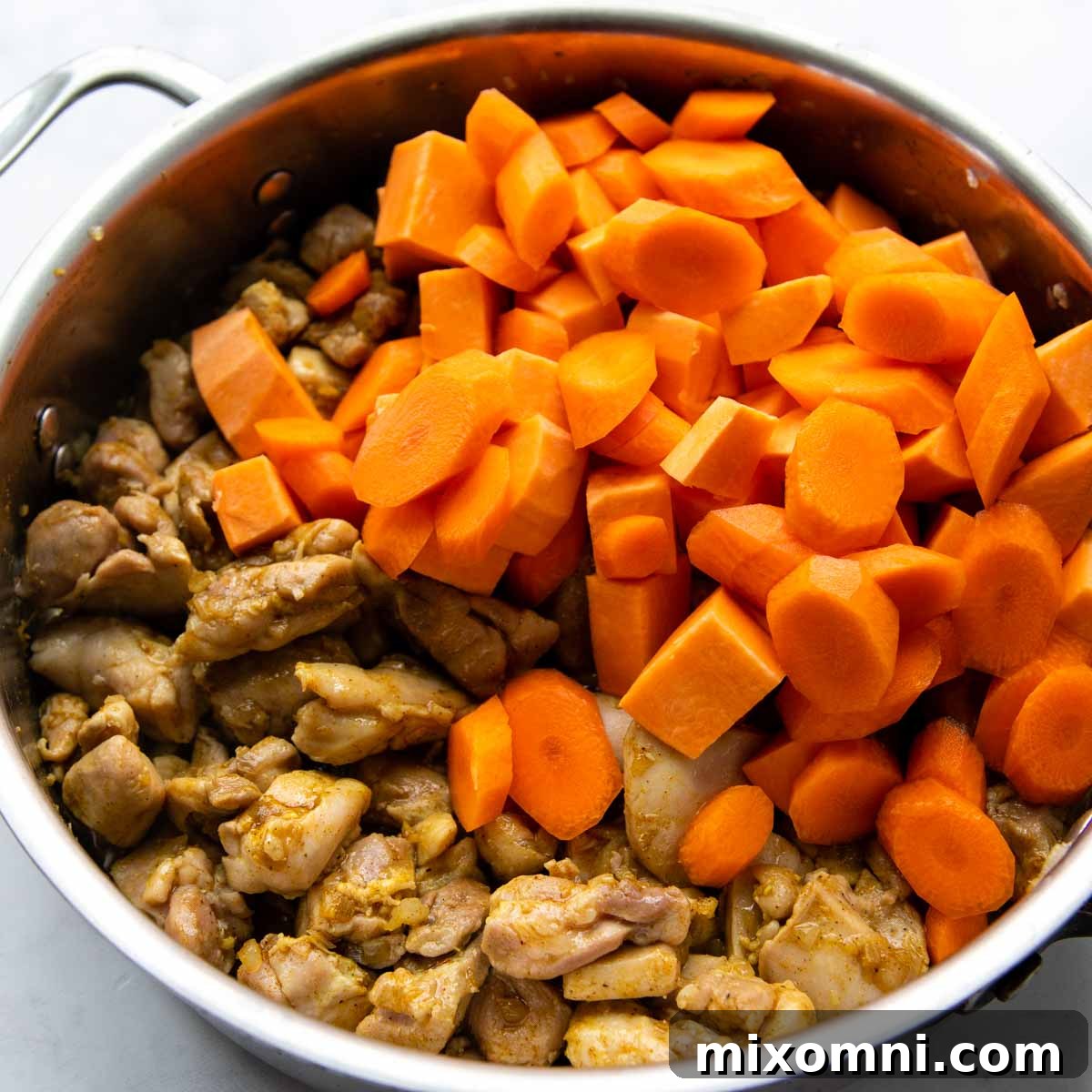 Carrots and sweet potatoes mixed with partially cooked chicken and aromatic spices in a skillet, ready for simmering.
