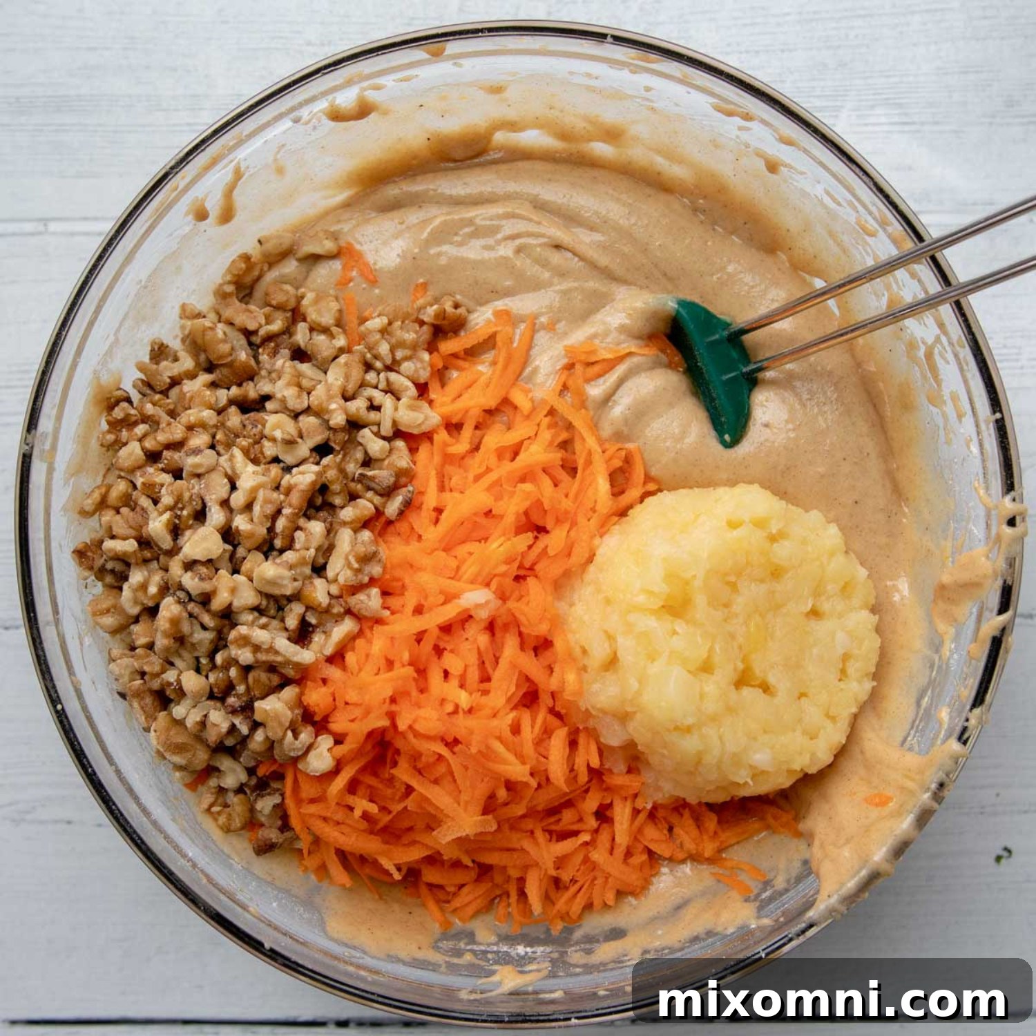 Grated carrots, crushed pineapple, and chopped nuts being stirred into the cake batter in a large bowl.