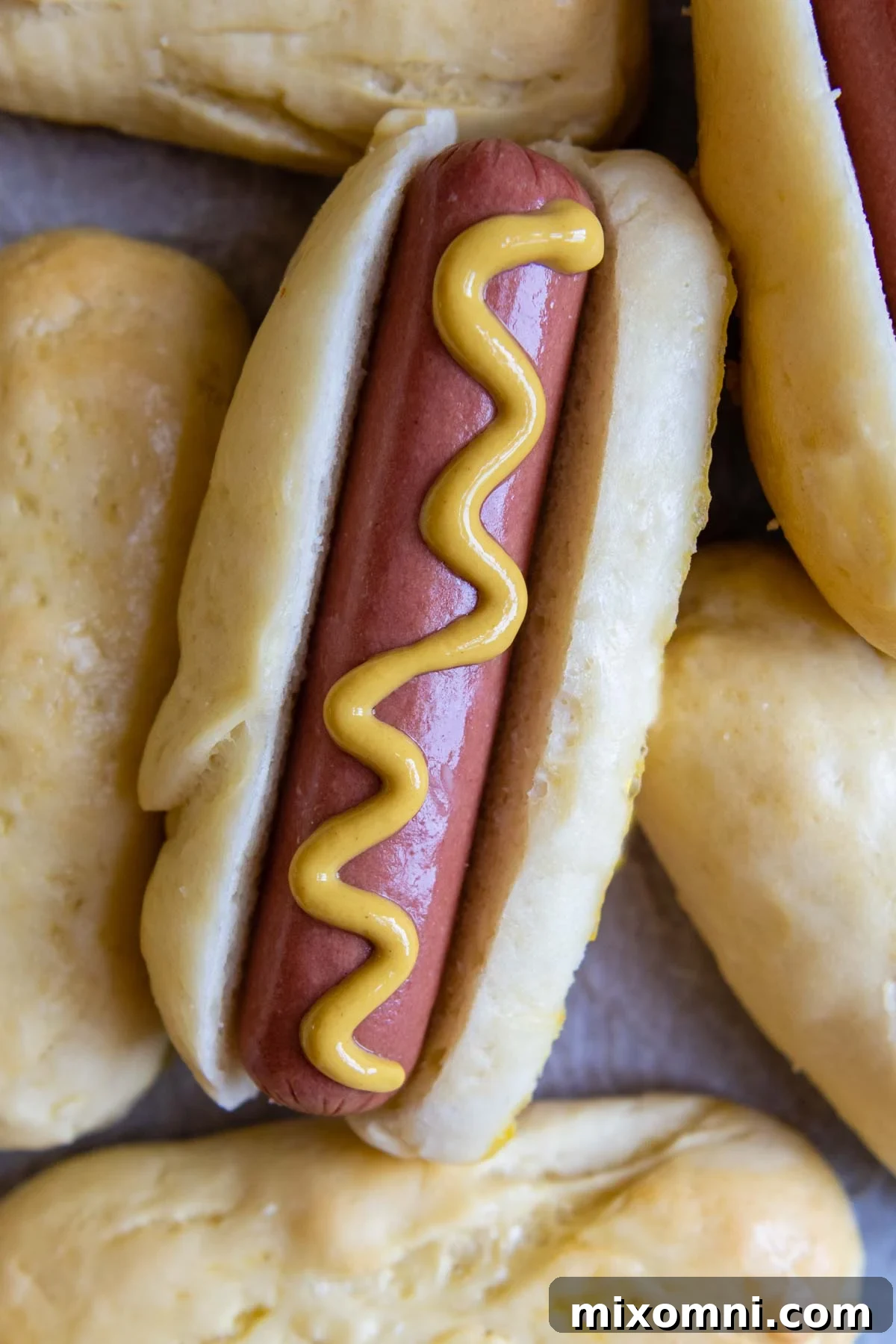 A perfectly cooked hot dog nestled in a soft, homemade gluten-free bun, drizzled with mustard. Other fluffy buns are visible in the background, ready to be enjoyed.