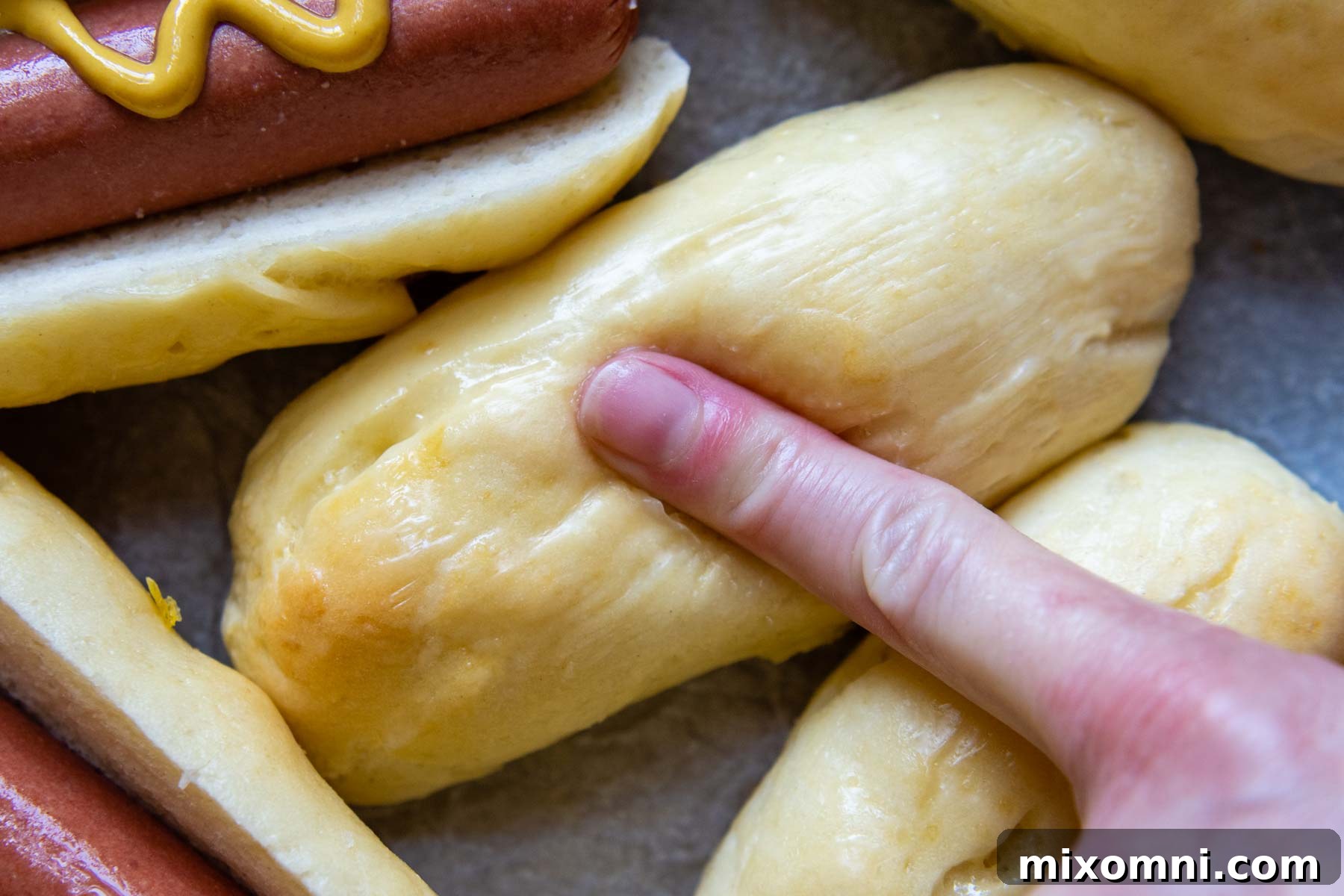 A finger gently pressing down on a fresh, soft gluten-free hot dog bun, demonstrating its squishy and resilient texture.