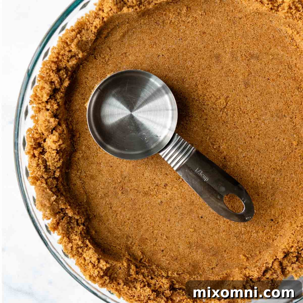 A measuring cup being used to firmly press the graham cracker mixture into the bottom of a pie plate, creating an even base.