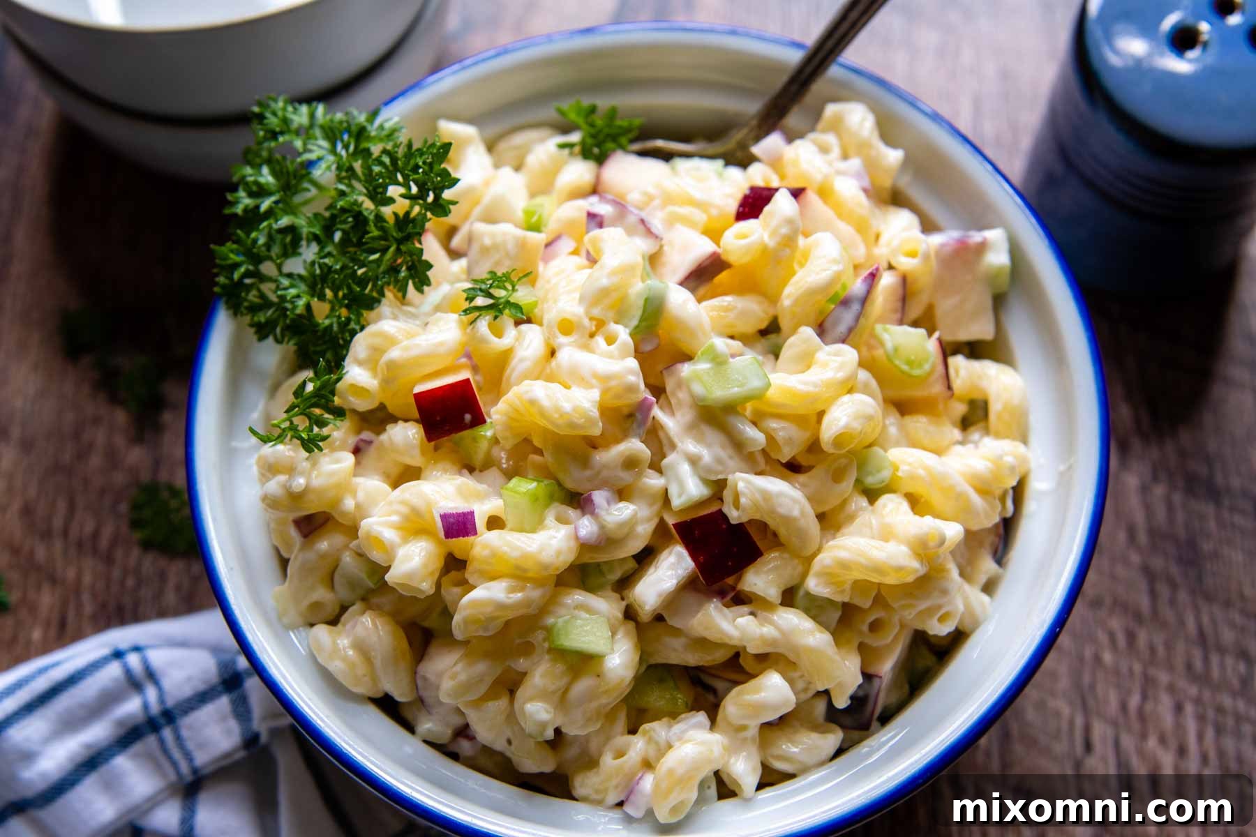 A white serving bowl of gluten-free macaroni salad, with salt and pepper shakers and a linen napkin beside it on a wooden surface.