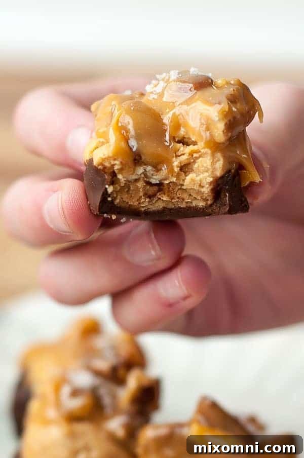 A hand holding up a buckeye-style peanut butter turtle with a bite taken out, revealing the creamy peanut butter and gooey caramel pecan layers.