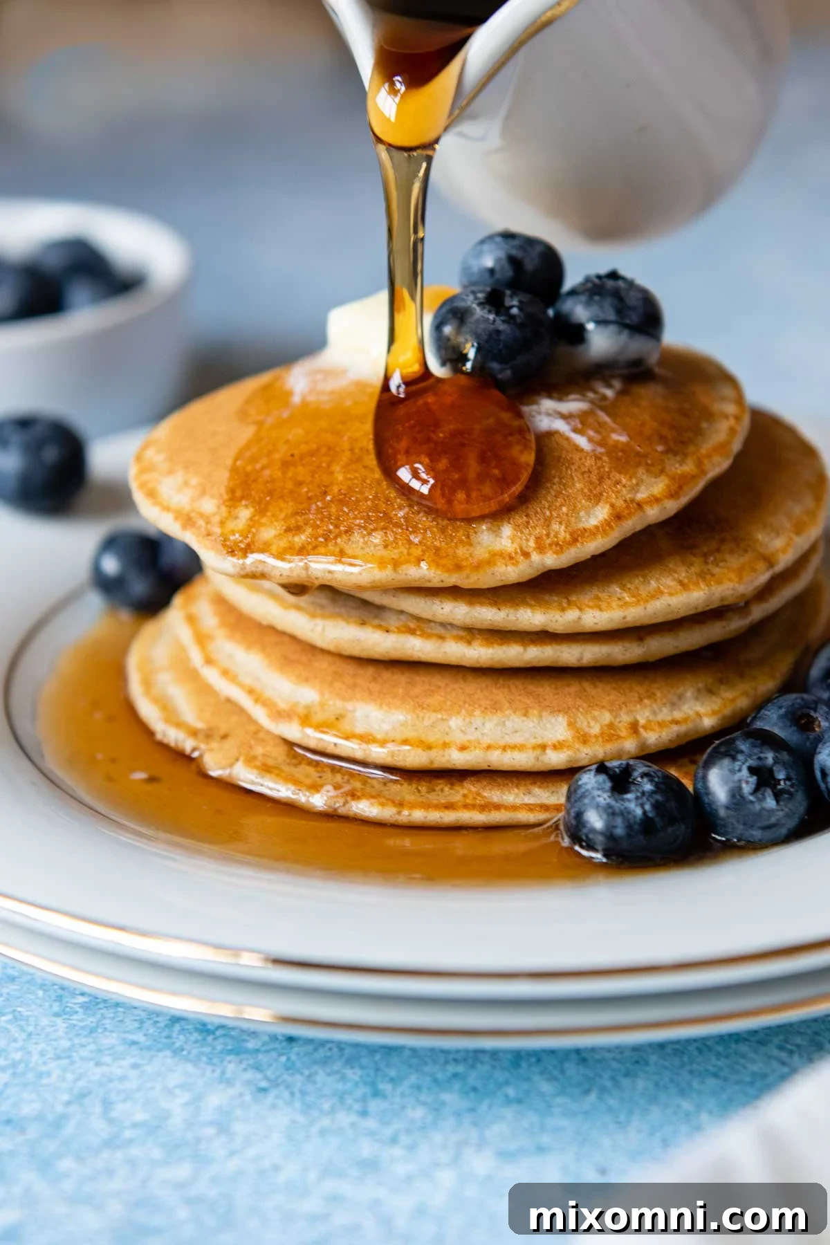 Golden syrup being generously poured over a tall, inviting stack of fluffy oat flour pancakes, garnished with fresh berries.