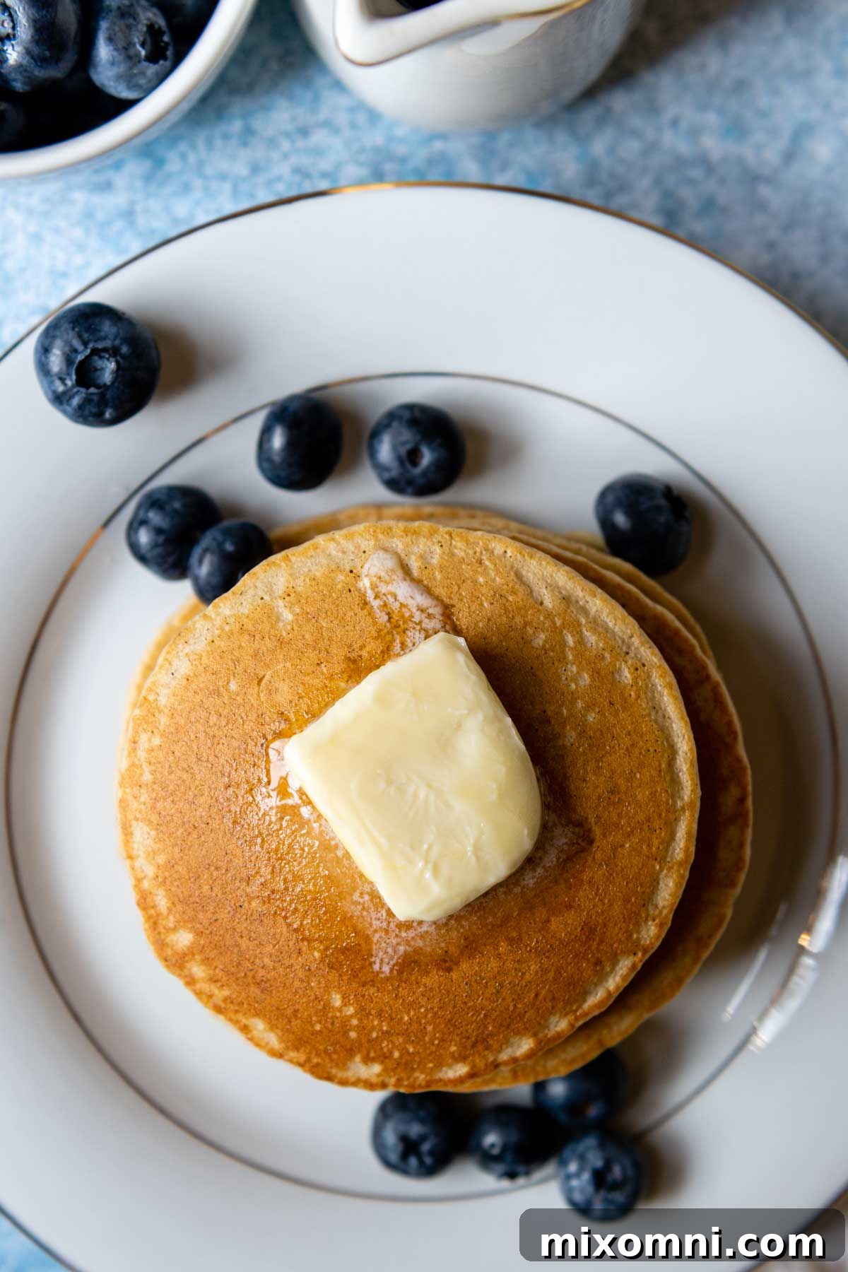 A beautiful stack of fluffy oat flour pancakes on a white plate, artfully scattered with fresh blueberries and powdered sugar.