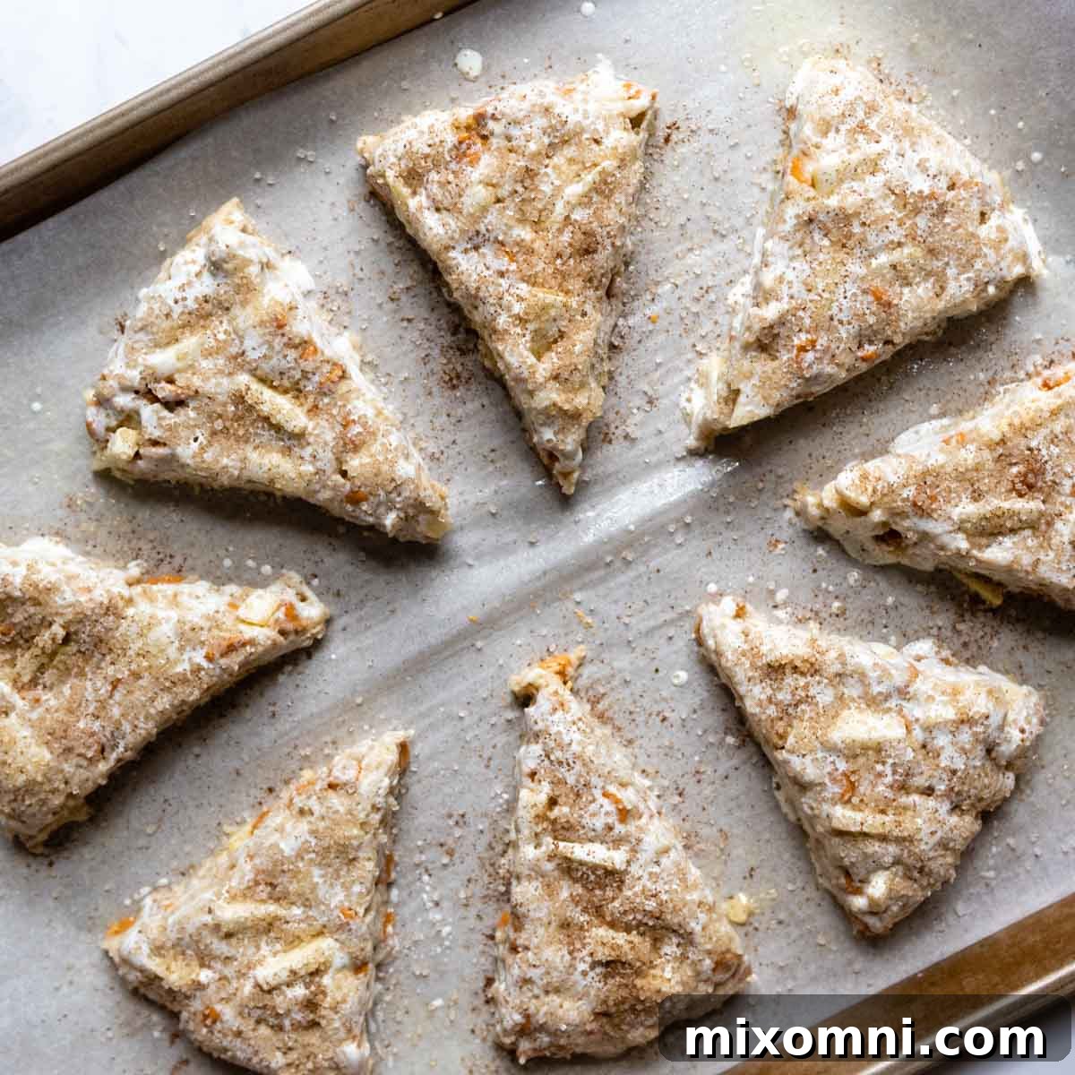 Unbaked scone triangles arranged on a baking sheet, brushed with milk and sprinkled with coarse sugar.
