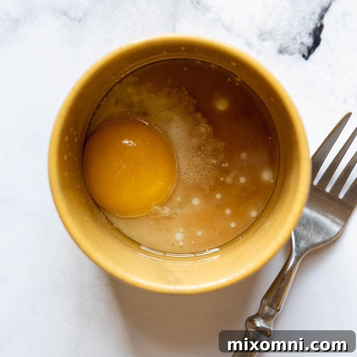 Liquid ingredients, including oil, milk, and vanilla extract, are whisked together with brown sugar in a mug, with a fork resting nearby.