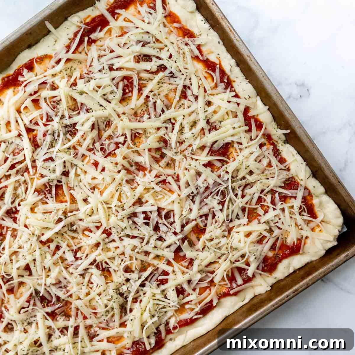 Pizza sauce and a generous layer of shredded cheese spread evenly over the gluten-free dough on a baking sheet.