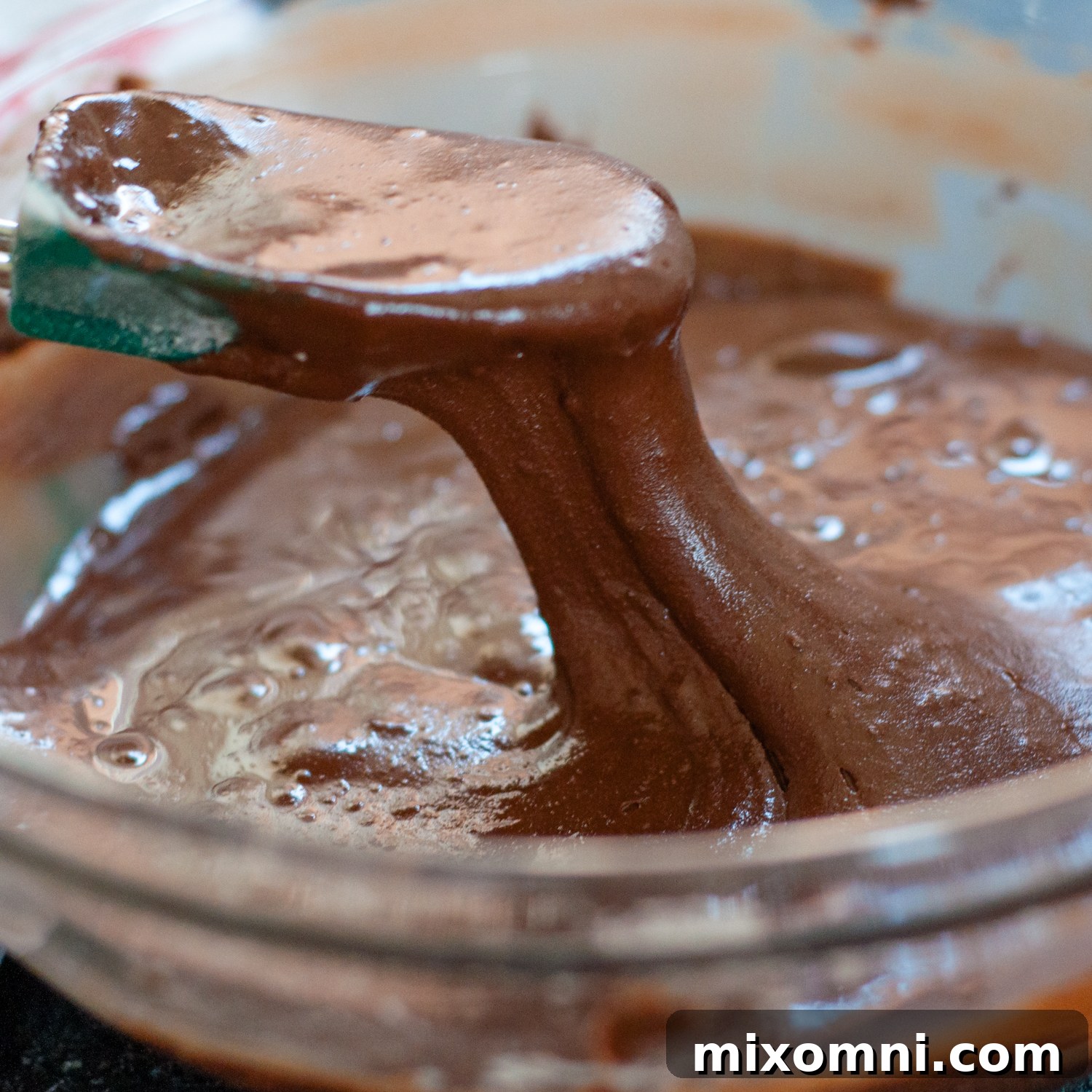 A silicone spoon stirring a very thick, dark chocolate cookie batter in a bowl, with chocolate chips visible.