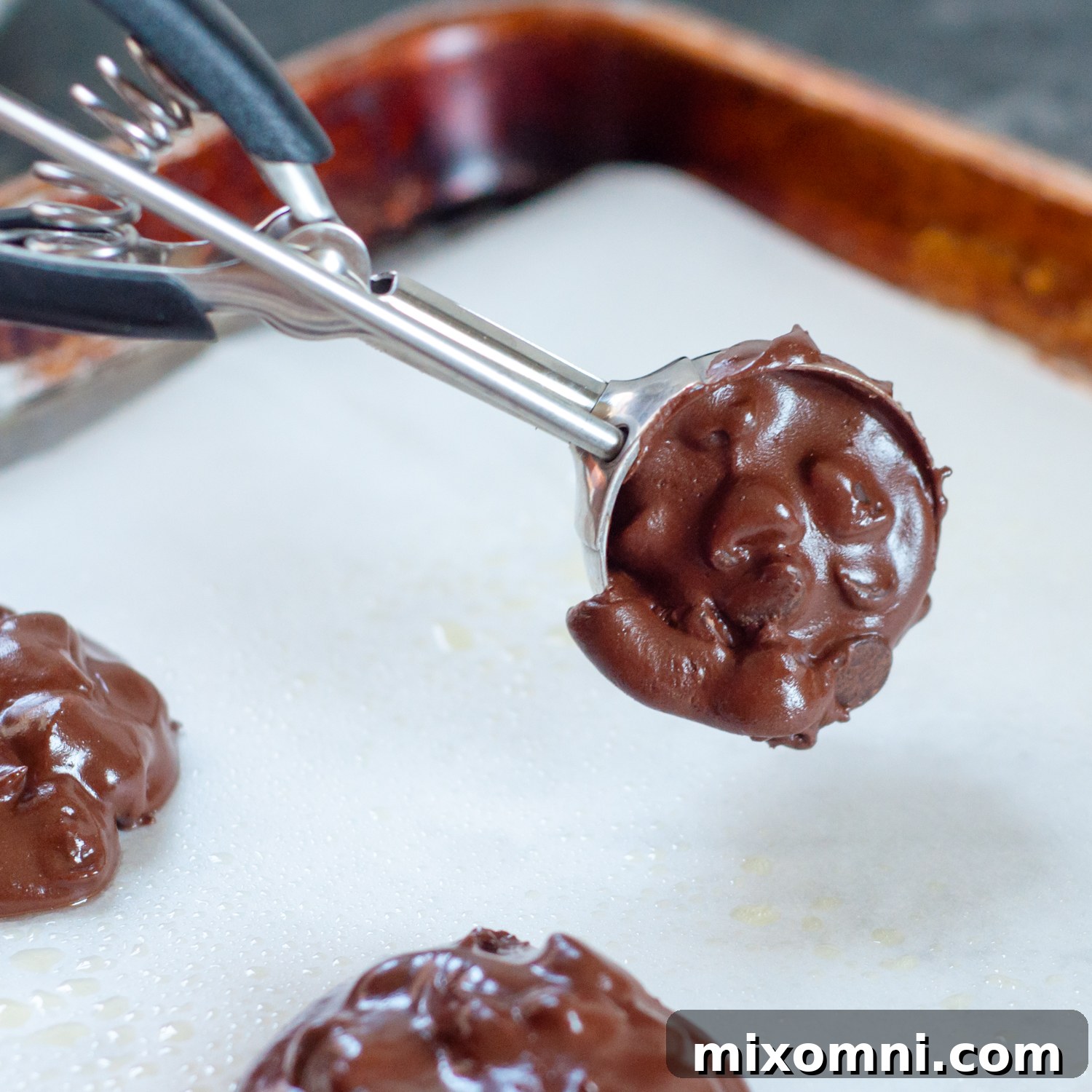 A small cookie scoop dropping portions of chocolate cookie batter onto a parchment-lined baking sheet.