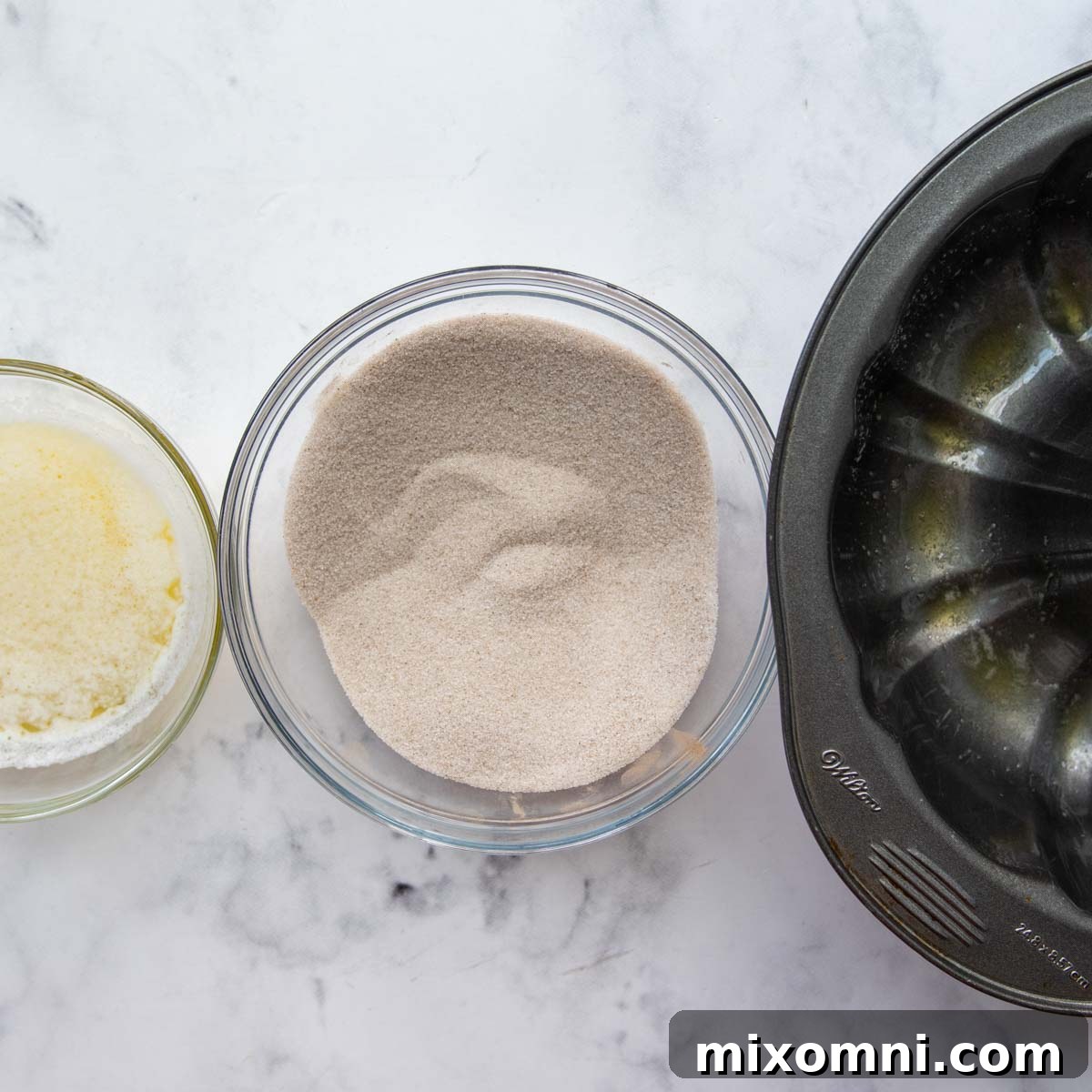 Greased bundt pan, a bowl of melted butter, and a bowl of cinnamon sugar mixture set up for the dipping station.
