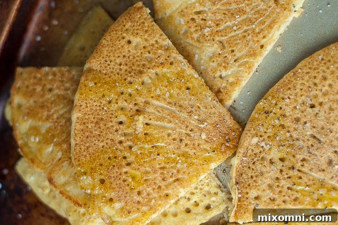 Socca cut into rustic, golden wedges, beautifully garnished with a generous drizzle of olive oil, coarse sea salt, and freshly ground black pepper, arranged on a textured surface.