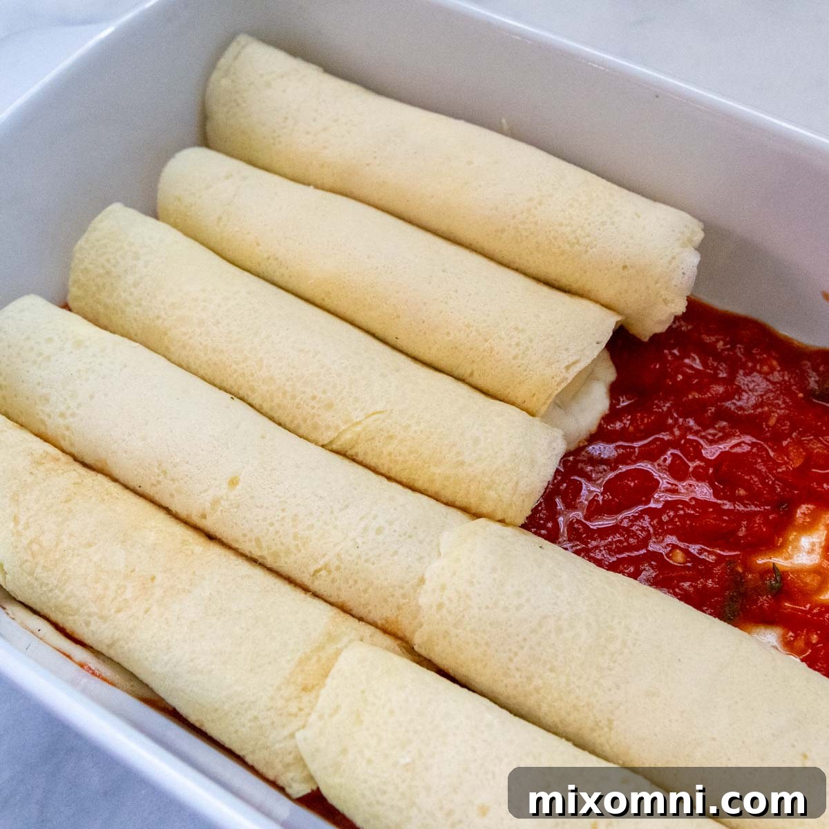 Unbaked gluten-free manicotti rolls neatly lined up in a casserole dish, topped with sauce.