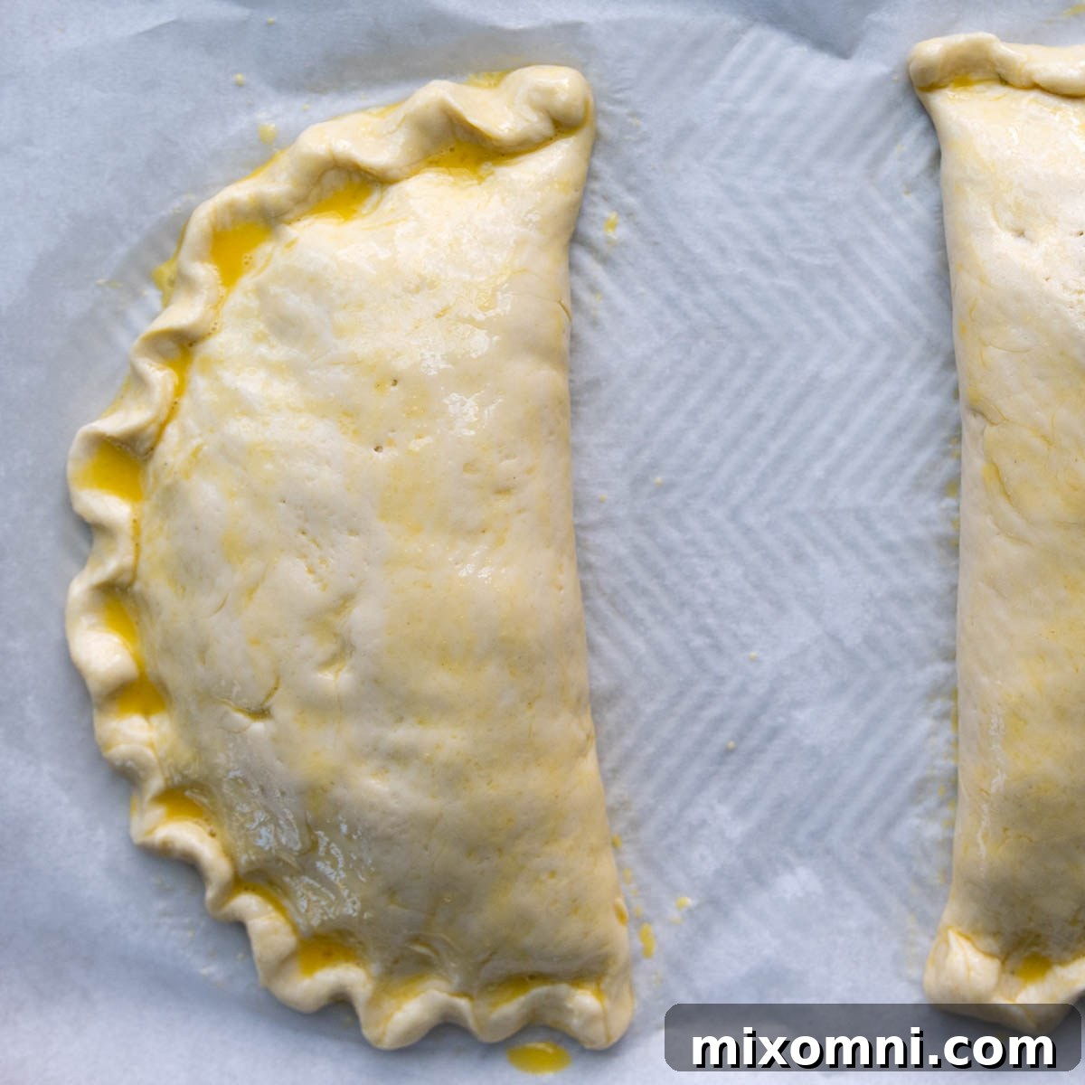 Two unbaked gluten-free calzones glistening with a fresh egg wash, ready to be placed in the oven for baking.