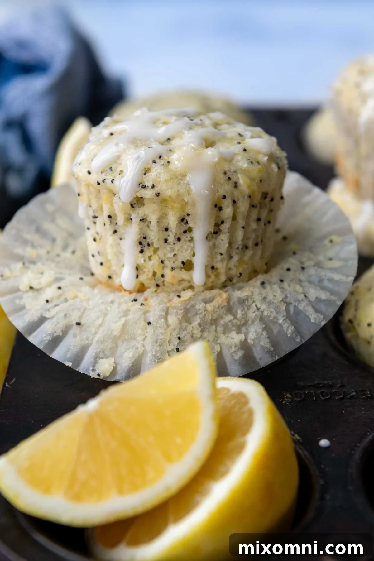 A golden-brown gluten-free lemon poppy seed muffin, partially unwrapped from its paper liner, with a bright white lemon glaze drizzled generously on top. The muffin reveals flecks of poppy seeds within its fluffy interior, suggesting a delightful texture and vibrant flavor.