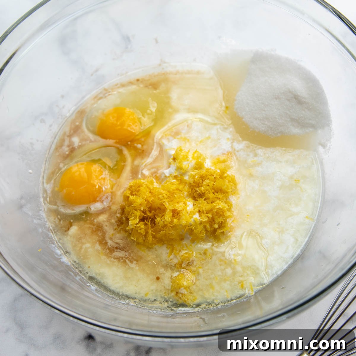 A clear glass mixing bowl holds a vibrant yellow mixture of wet ingredients including melted butter, oil, granulated sugar, sour cream, eggs, lemon juice, lemon zest, and extracts, ready for dry ingredients.