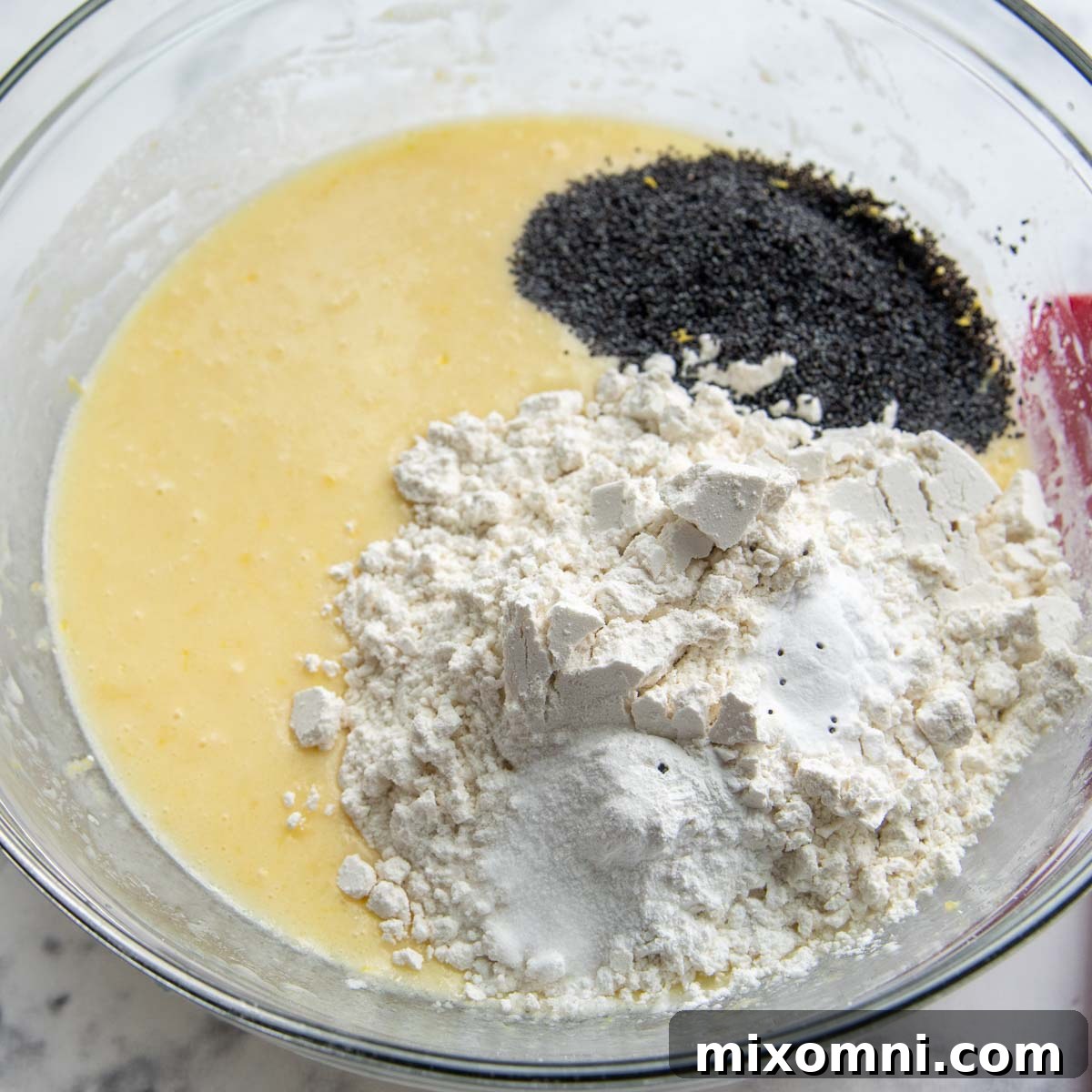 A glass bowl containing the mixed wet ingredients, with a layer of dry ingredients including gluten-free flour and poppy seeds poured on top, ready to be gently stirred together to form the muffin batter.