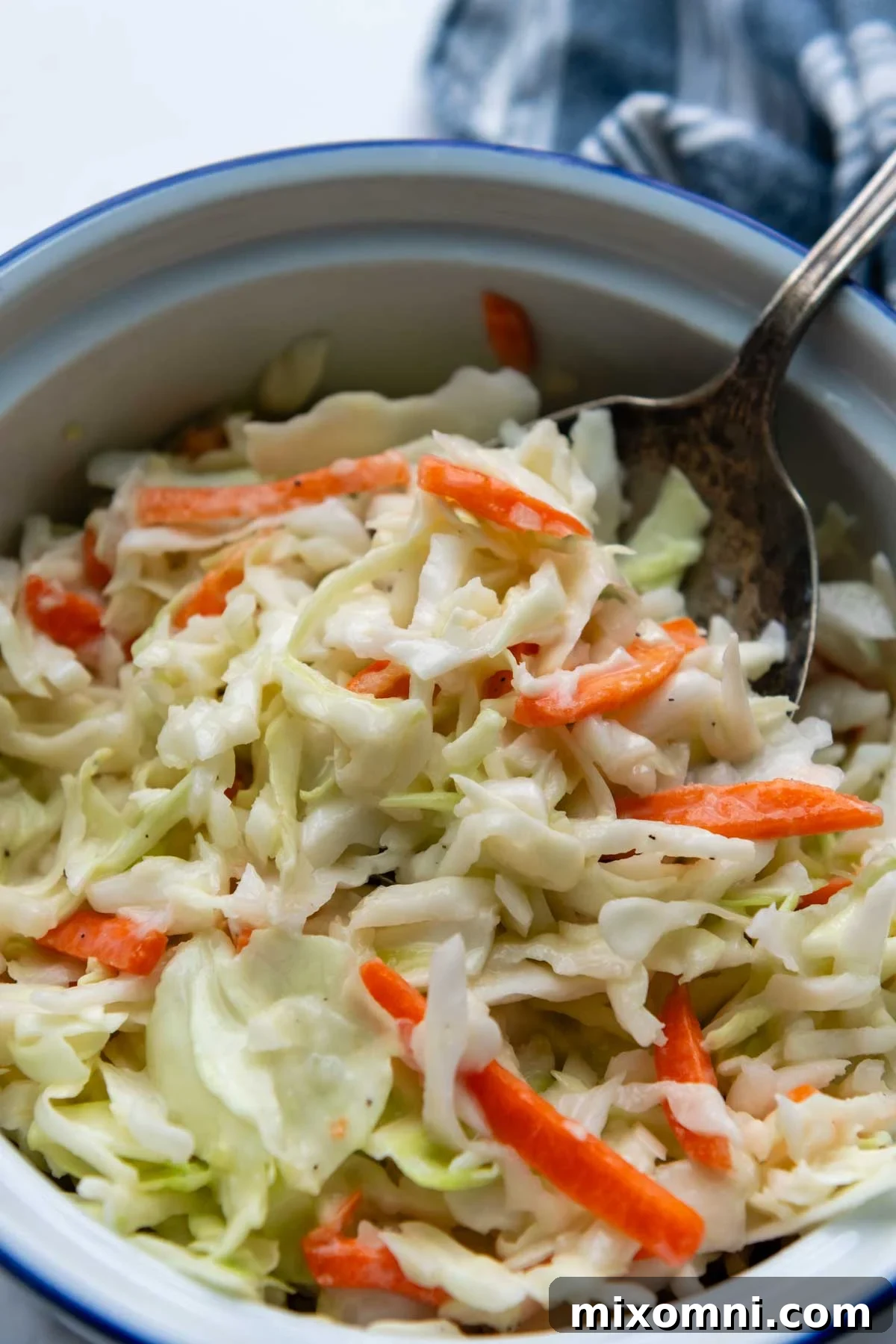 A spoon going into a bowl of creamy gluten-free coleslaw, presented beautifully on a white marble surface.