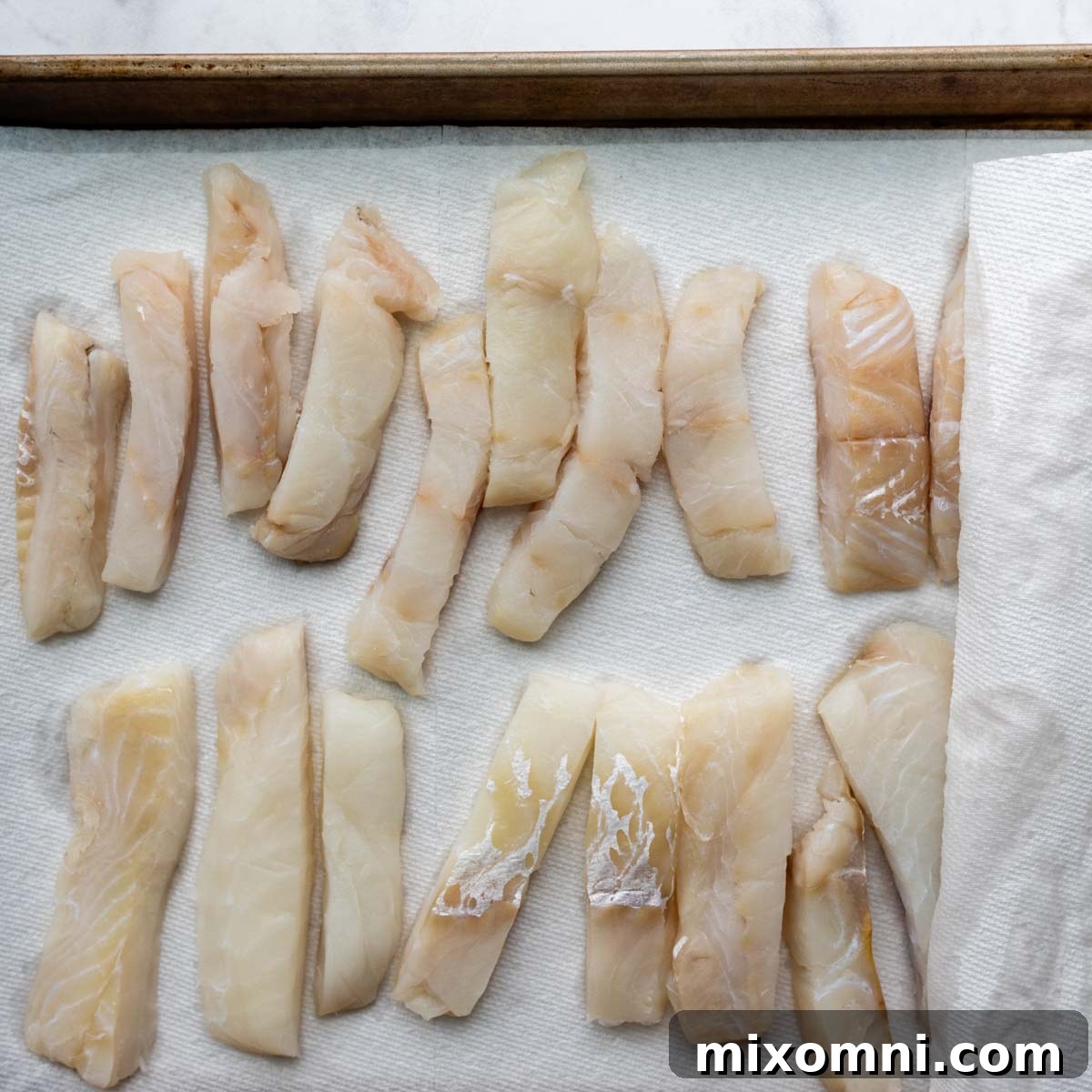 Carefully arranged pieces of white fish fillets on a baking sheet, lined with absorbent paper towels to remove excess moisture before dredging.