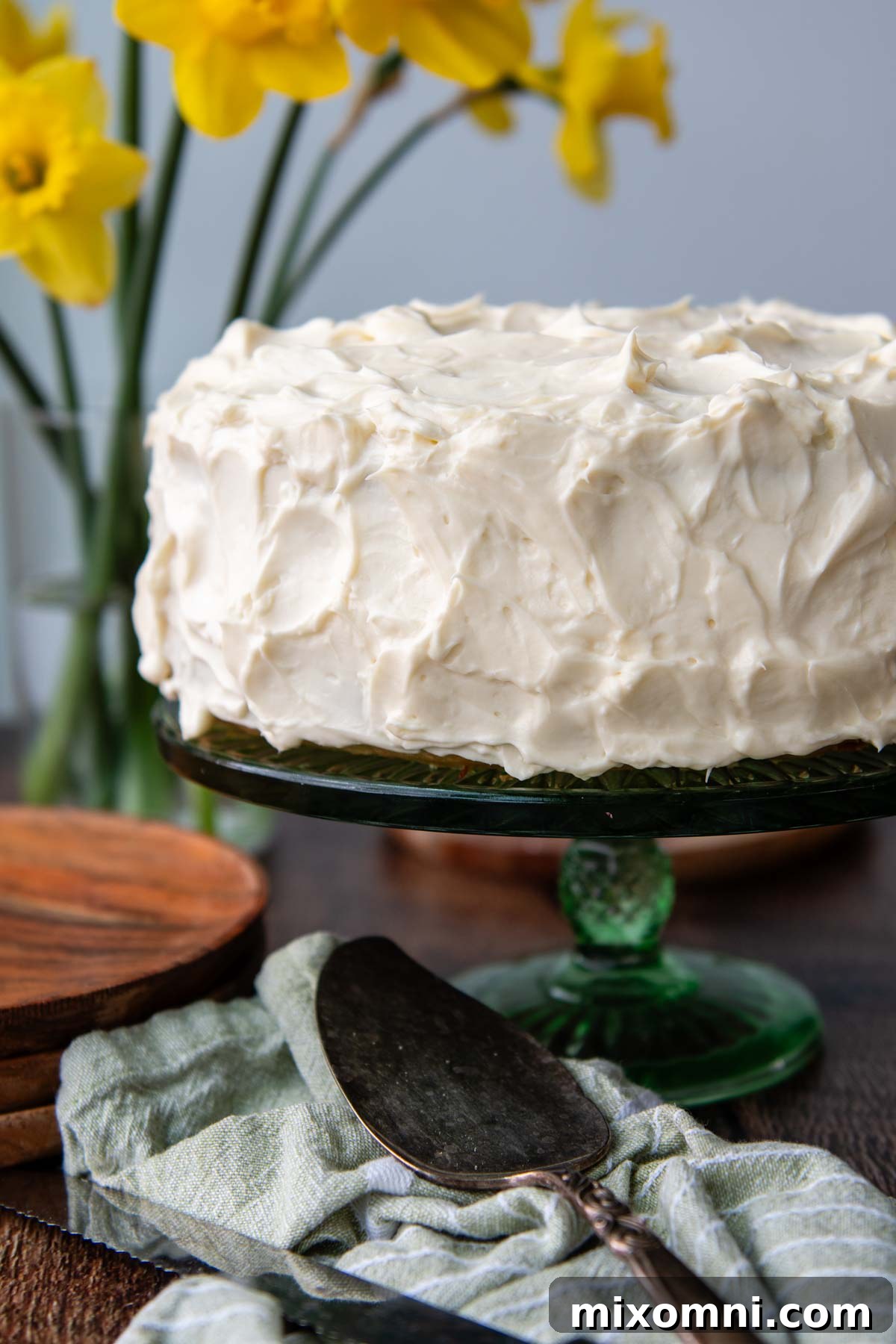 A beautifully frosted whole gluten-free banana cake on a vibrant green platter, surrounded by fresh flowers.