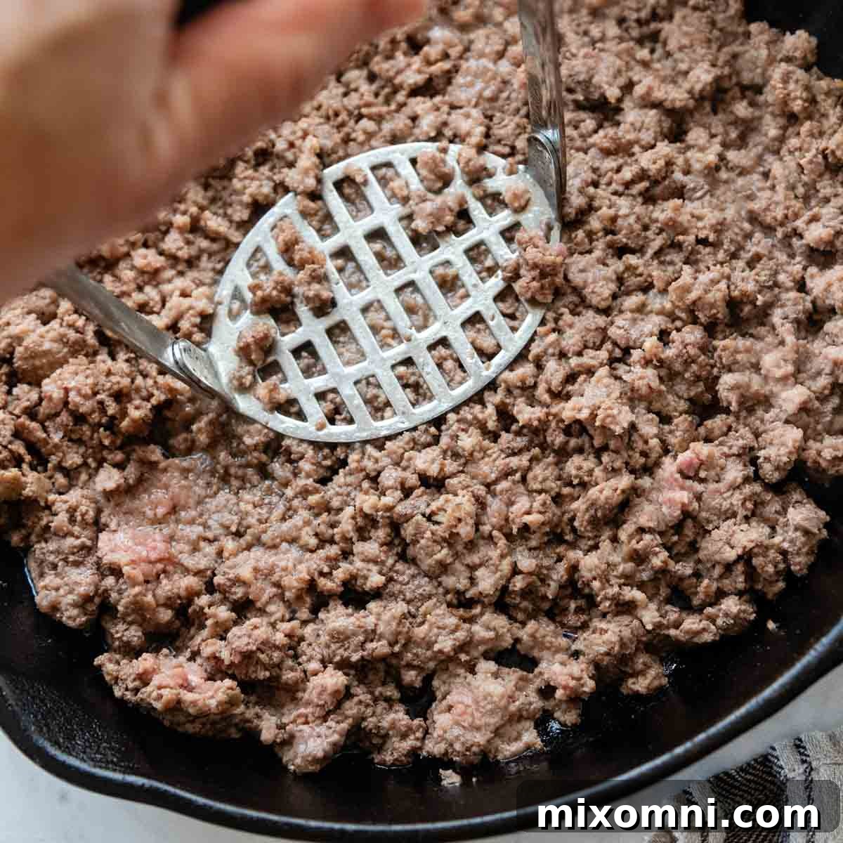 A potato masher actively breaking up browned ground beef in a skillet, creating a fine, even texture.