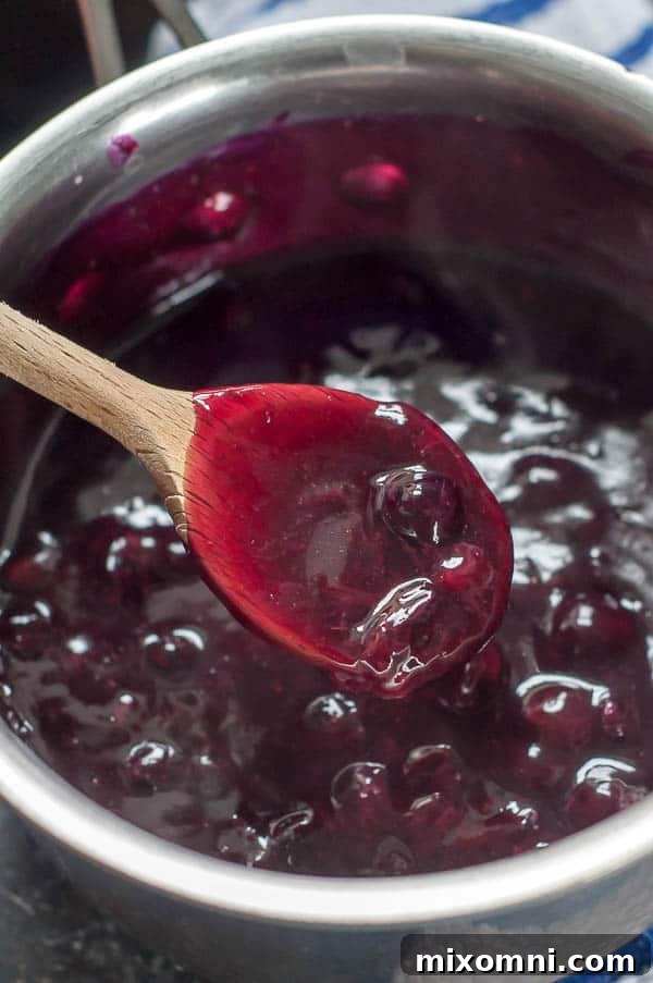 A wooden spoon holding up thickened blueberry syrup from a saucepan, demonstrating the desired consistency.