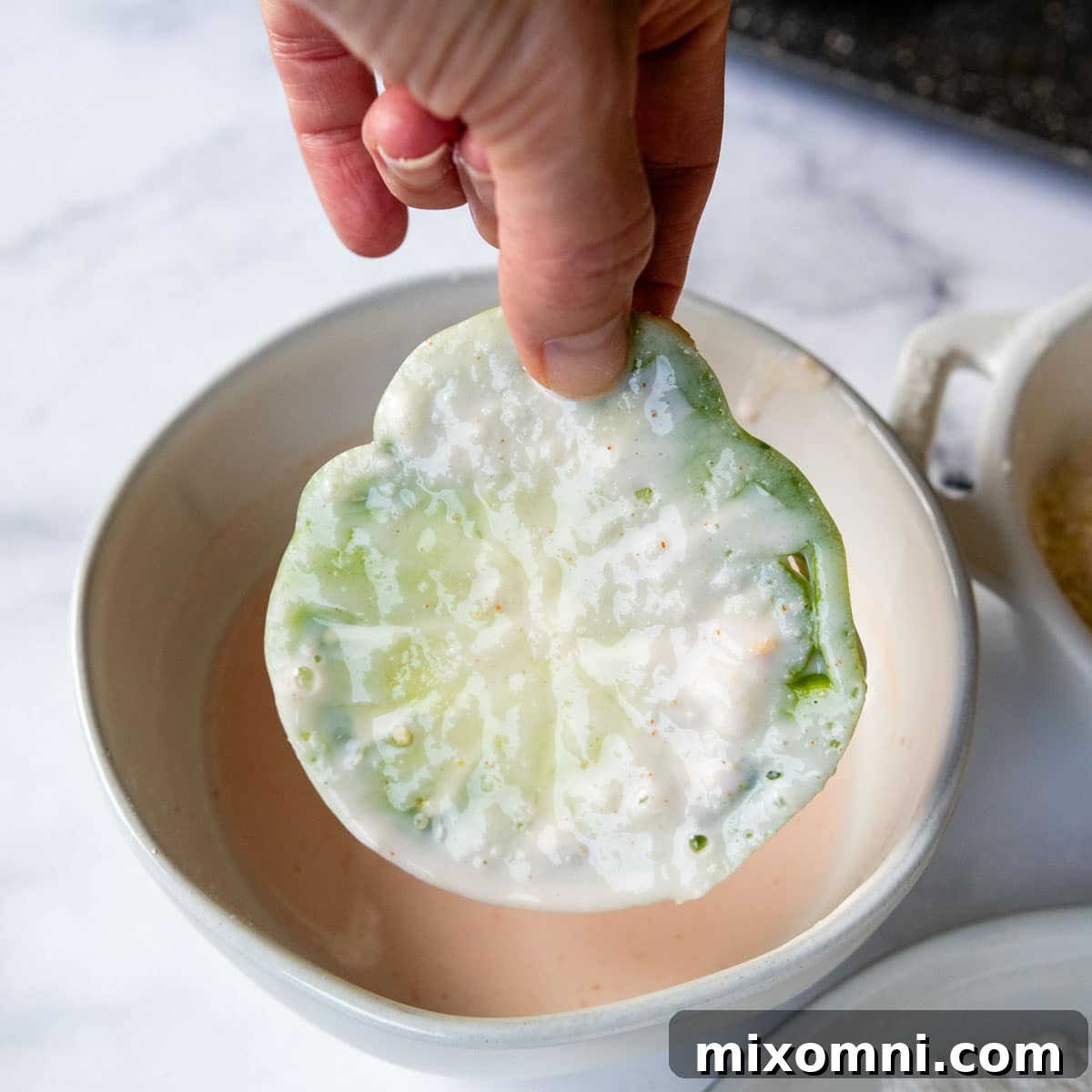 A green tomato slice being lifted from the buttermilk mixture, excess dripping off.