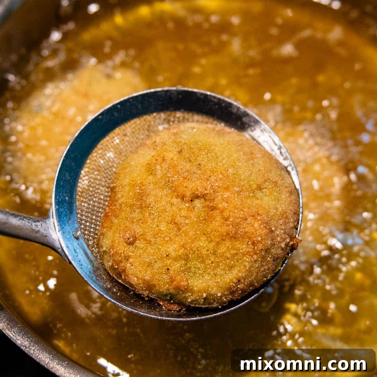 A skimmer spoon carefully lifting a golden-brown fried green tomato out of hot oil.