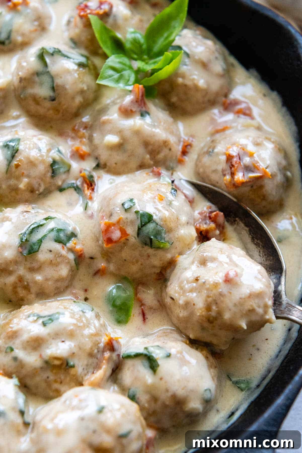 A spoon lifting a juicy gluten-free chicken meatball out of a pan filled with creamy sun-dried tomato sauce, ready to be served.