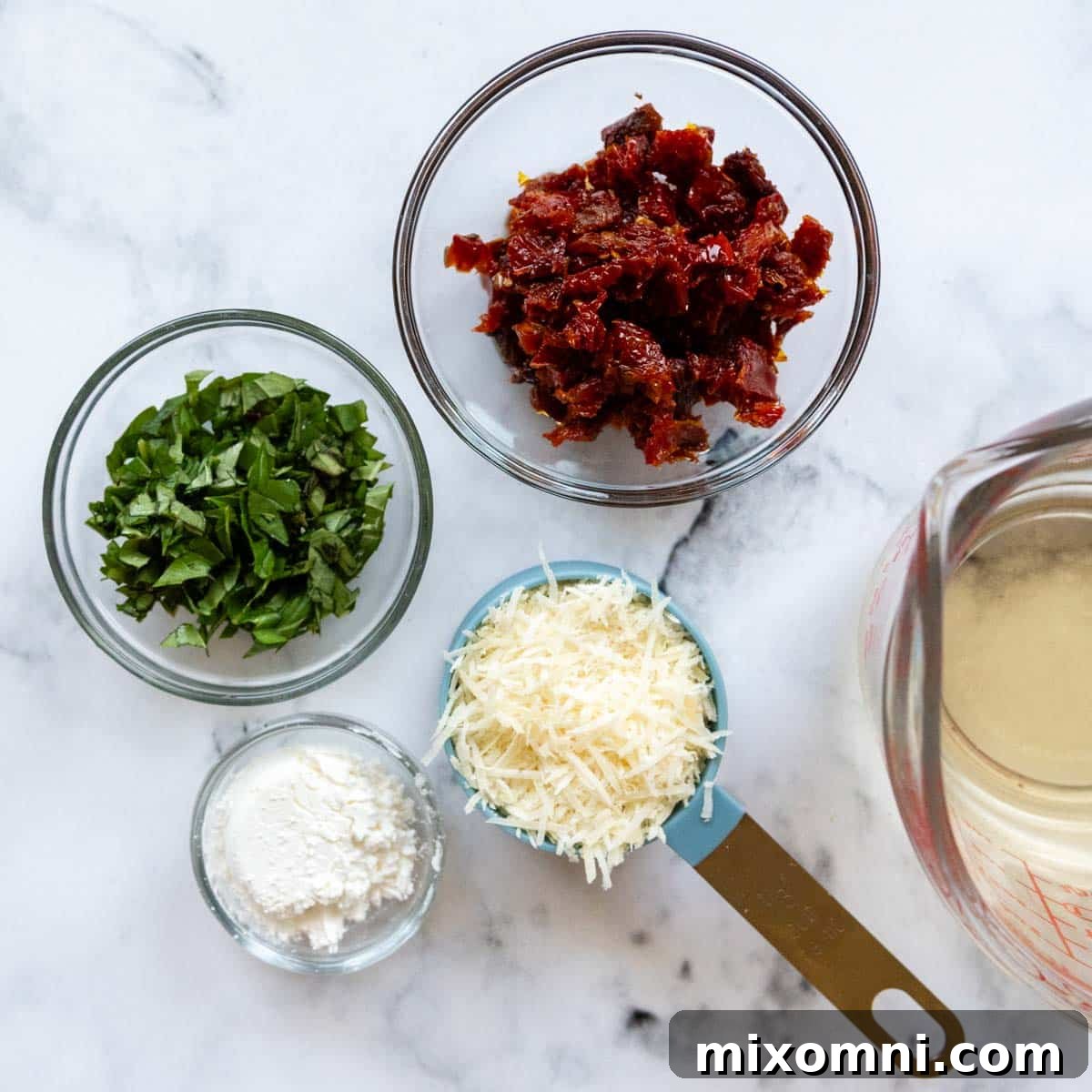 A collection of fresh ingredients for the creamy sun-dried tomato sauce, including minced garlic, chopped basil, grated parmesan, and sun-dried tomatoes in oil.