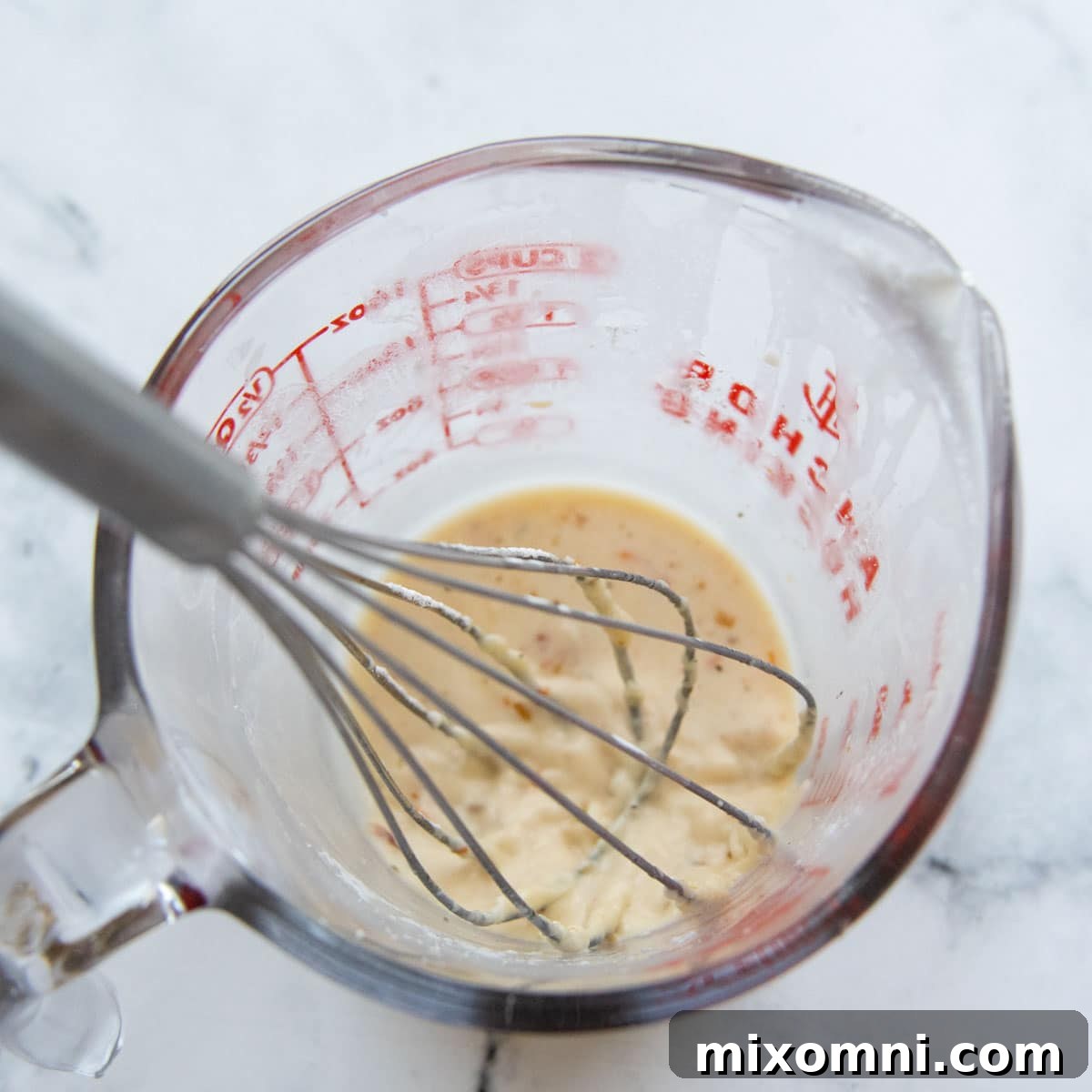 A whisk being used in a liquid measuring cup to combine cornstarch with a small amount of hot sauce, creating a thickening slurry.