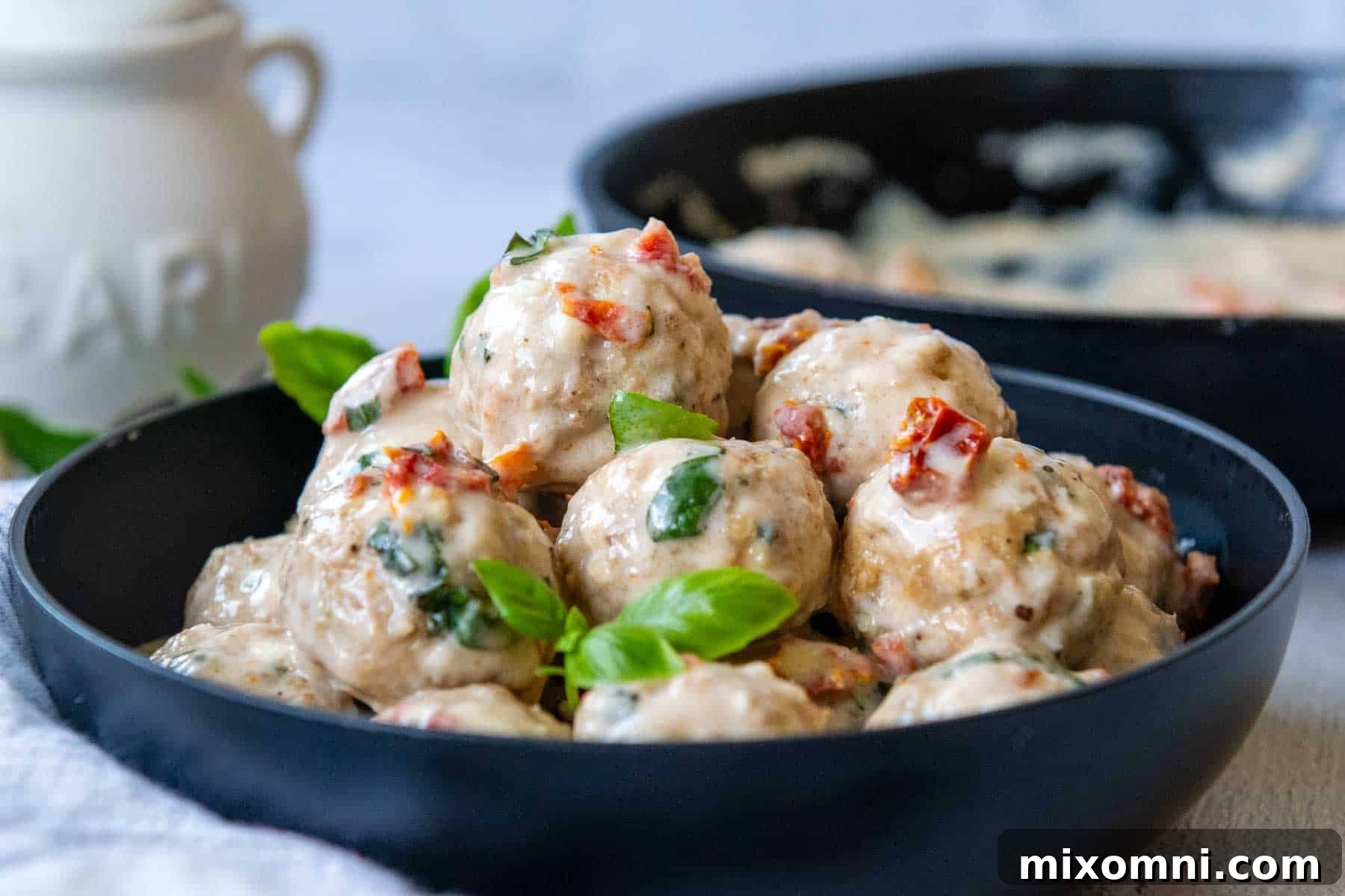 A shallow black bowl overflowing with perfectly cooked gluten-free chicken meatballs drenched in creamy sun-dried tomato sauce, garnished with fresh basil leaves.