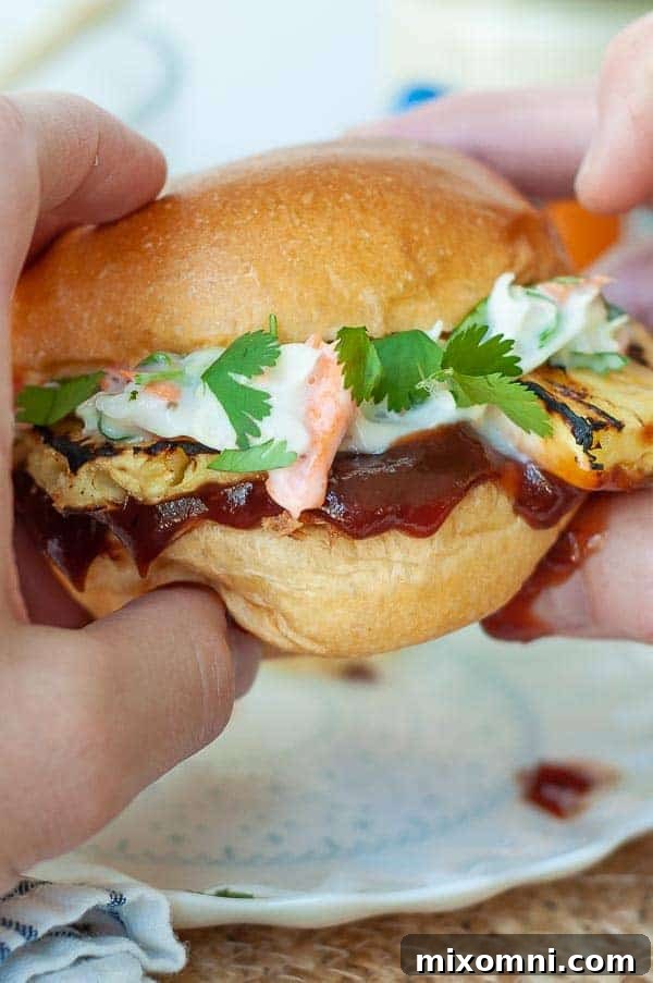 Two hands holding up a generously loaded BBQ pulled chicken sandwich, showcasing the tender chicken, grilled pineapple, and creamy slaw.