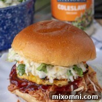 A BBQ pulled chicken sandwich on a plate with coleslaw, BBQ sauce, and grilled pineapple