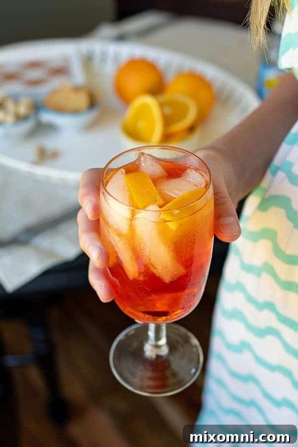 A hand holding a vibrant orange Aperol Spritz cocktail in a large wine glass, garnished with a fresh orange slice, against a soft, blurred background.